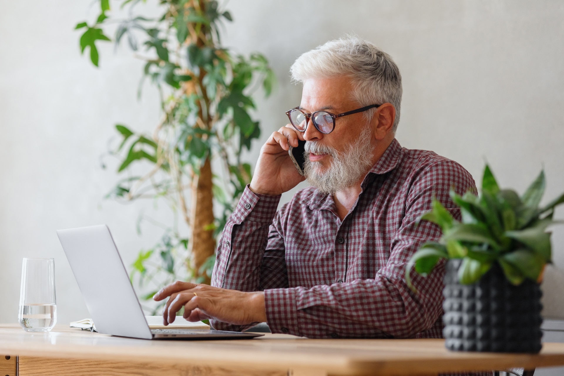 Un hombre de pelo gris trabajando en una oficina con un ordenador portátil y hablando por teléfono. Un hombre adulto director o empresario negociando en una cámara web online. 