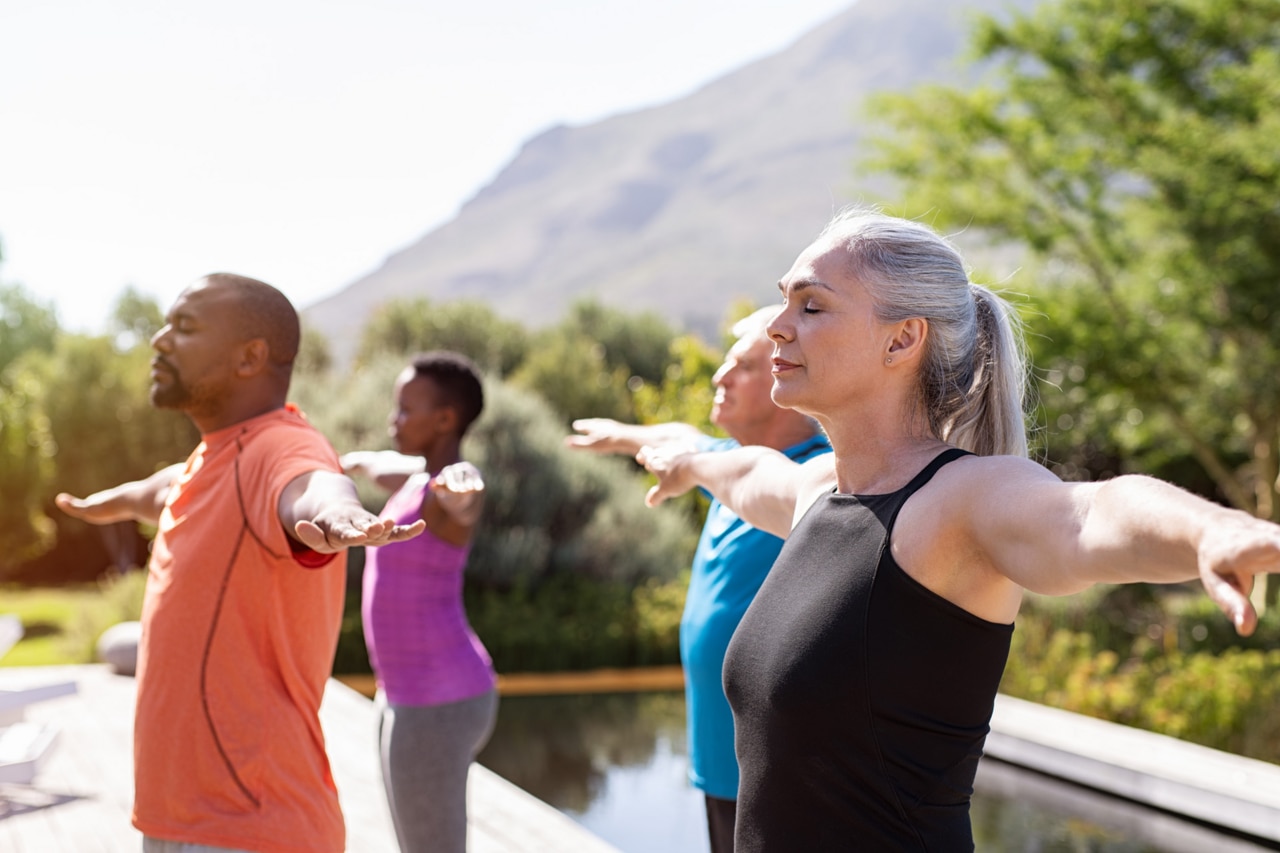 Gruppe älterer Menschen streckt draußen mit geschlossenen Augen die Arme. Atemübungen während Yogakurs für Senioren. Frauen und Männer machen Atemübungen mit ausgestreckten Armen.