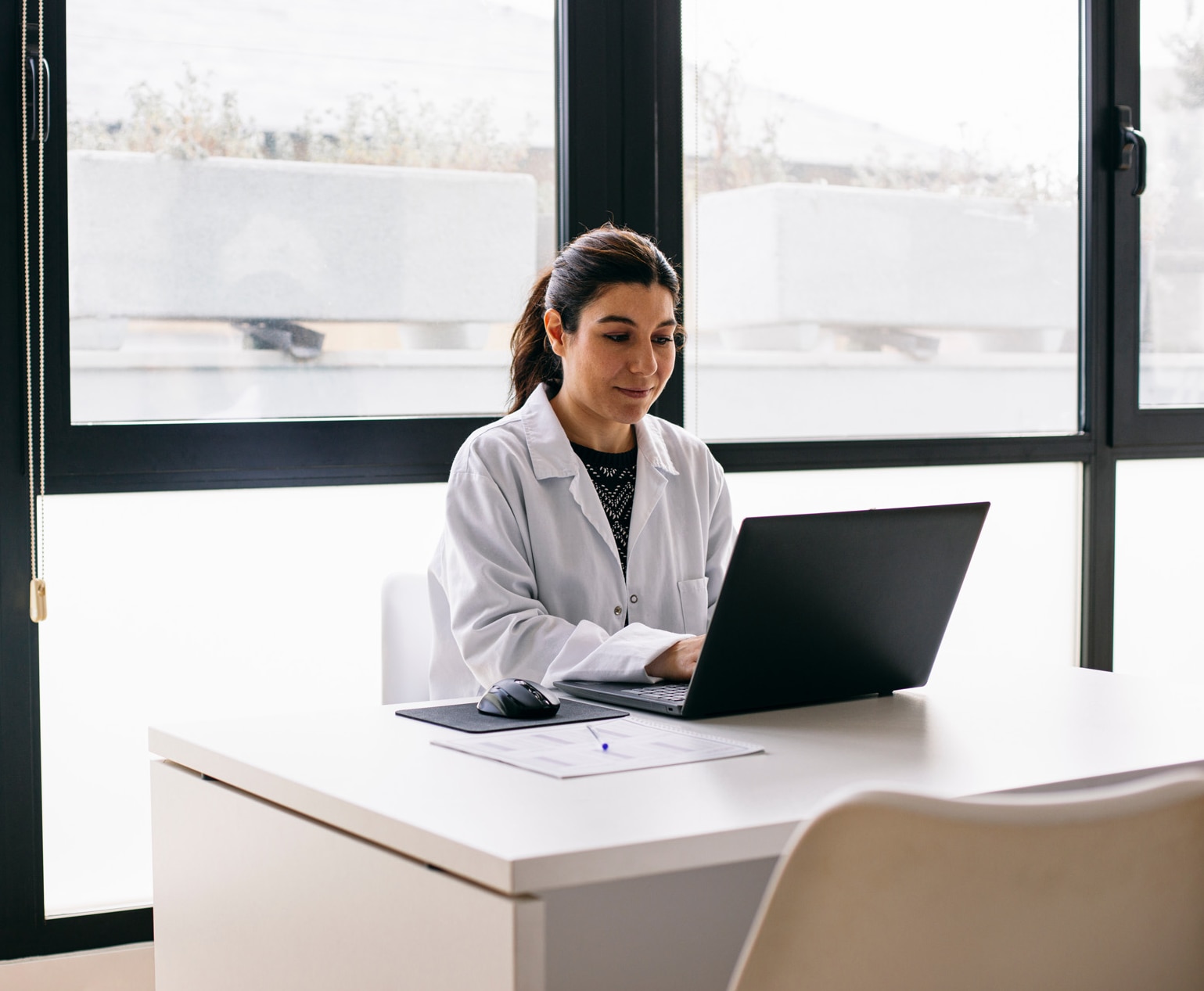 Kvindelig læge sidder ved et skrivebord i praksissen og bruger en laptop.