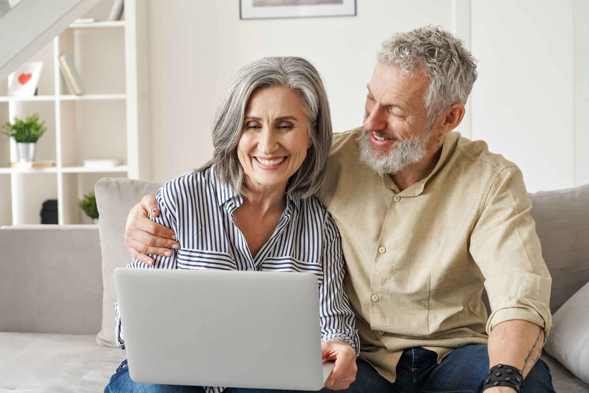 Et glad midaldrende par bruger en laptop siddende på sofaen, hvor de handler online på et website. Smilende ældre mand og kvinde ser på en computer fra deres hjem, hvor de bestiller. forsikringer på internettet.