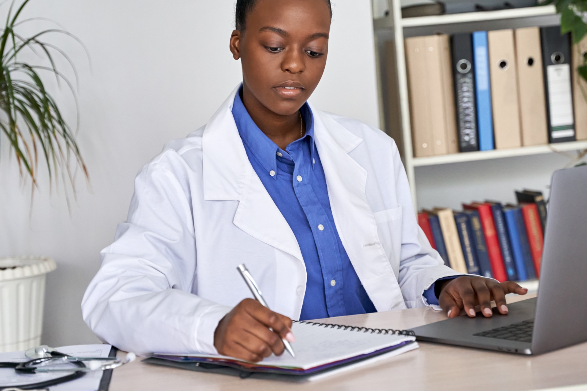 Afroamerikkalainen yleislääkäri valkoisessa takissaan ja stetoskoopissaan kirjoittamassa potilaskertomusta tietokoneellaan, selaamassa internetiä tai katsomassa webinaaria työpöytänsä ääressä