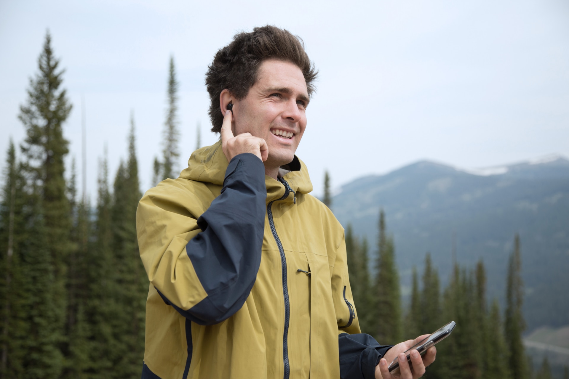 Un uomo che indossa un apparecchio acustico Virto Marvel con uno smartphone in mano conversa in un paesaggio di montagna.
