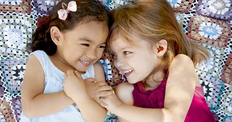 Två glada flickor i fyraårsåldern – en av dem har en röd hörapparat i sitt vänstra öra.