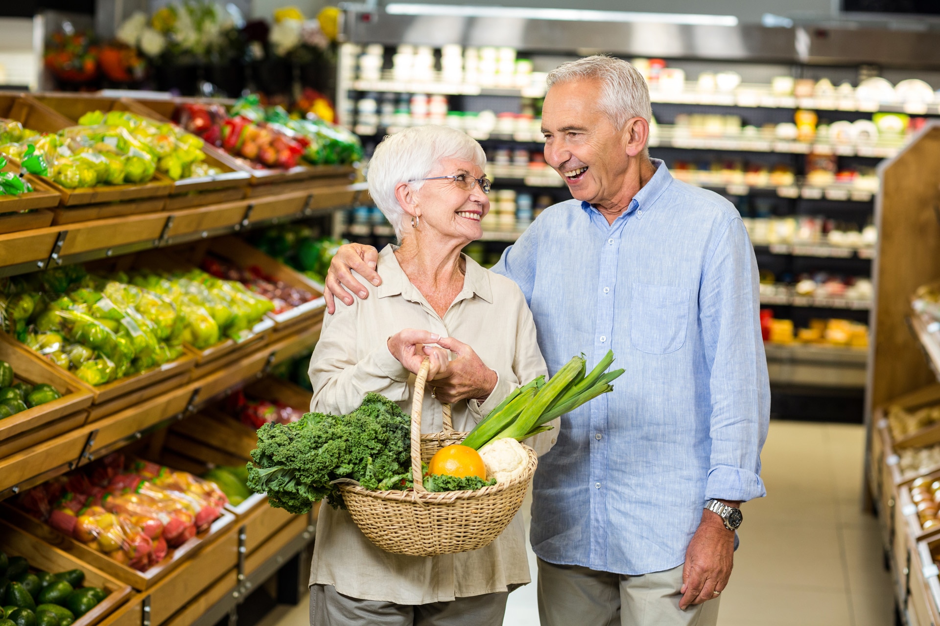 Leende äldre par som håller i en korg med grönsaker i en livsmedelsbutik.