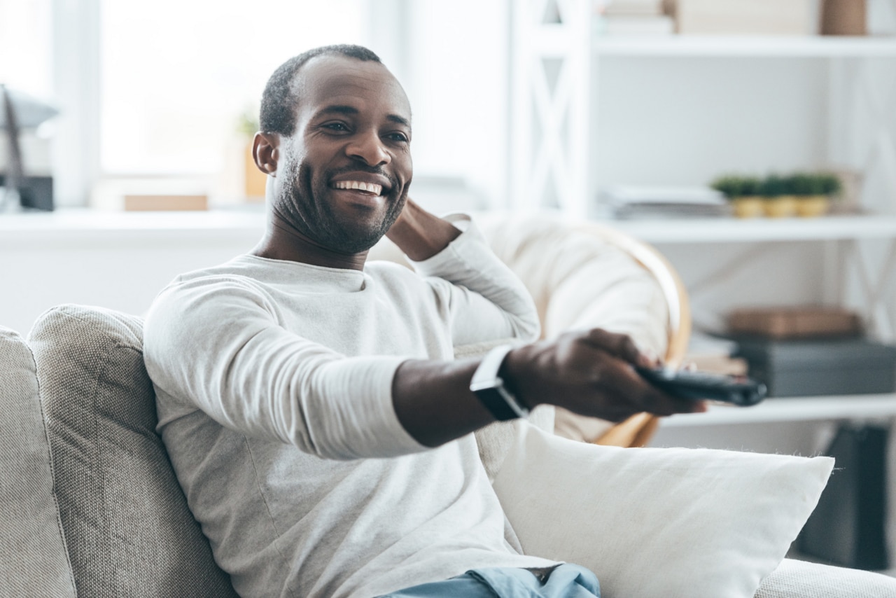 TV-tittande i hemmet. En ung, stilig afrikansk man sitter hemma i sin soffa och tittar på TV och ler