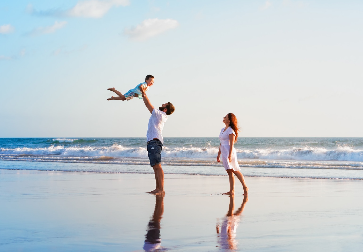Happy family holidays. Joyful father, mother, baby son walk with fun along edge of sunset sea surf on black sand beach.