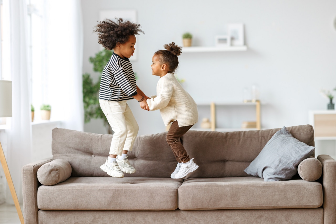 Carefree childhood. Happy energetic african american children jumping on sofa while playing game together at home