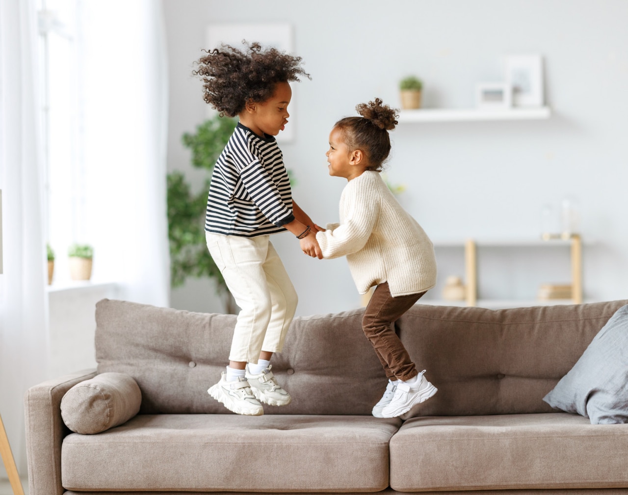 Carefree childhood. Happy energetic african american children jumping on sofa while playing game together at home, small active kids brother and sister having fun in living room; Shutterstock ID 2060068835; purchase_order: -; job: -; client: -; other: -