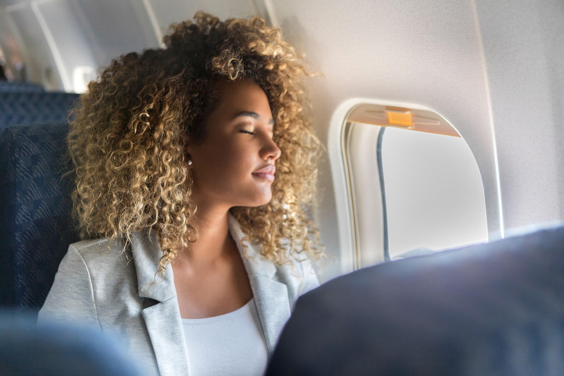 Eine junge Frau sitzt am Fensterplatz eines Linienflugzeugs und lehnt sich mit geschlossenen Augen zurück.