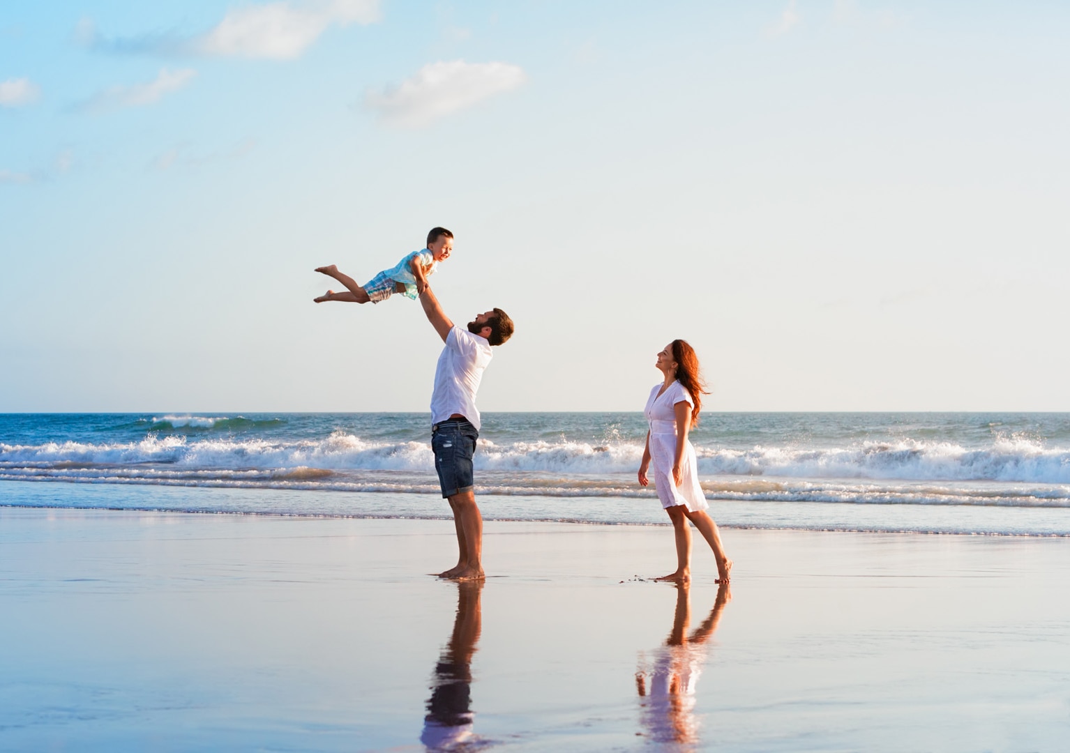 Happy family holidays. Joyful father, mother, baby son walk with fun along edge of sunset sea surf on black sand beach.