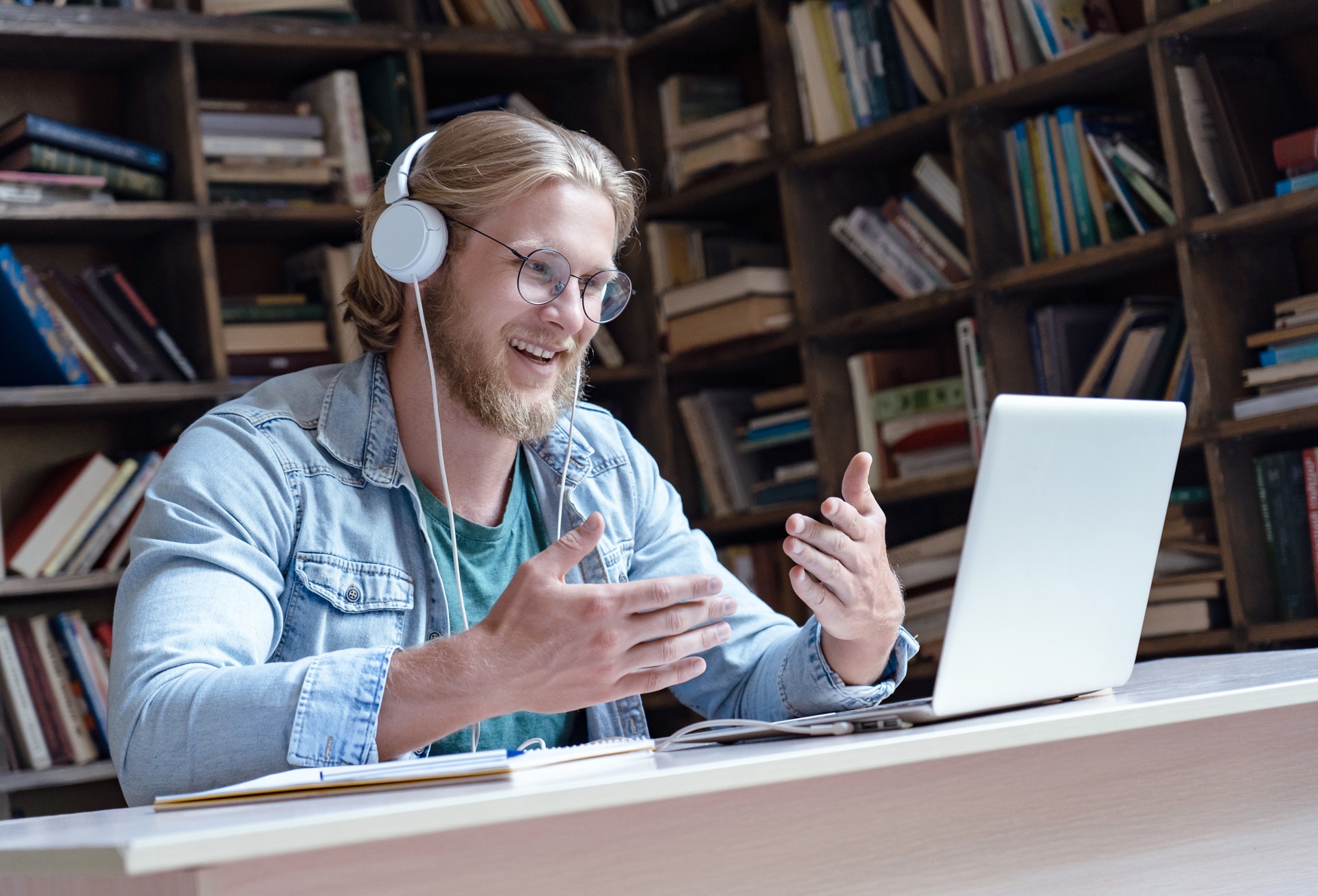 Lächelnder Student online, Lehrer trägt Kopfhörer, sprechen, Videoanruf, schaut auf den Bildschirm eines Laptops, kommuniziert über einen Chat mit einem Skype-Tutor, Fernstudium per E-Learning-Kurs in Bibliothek