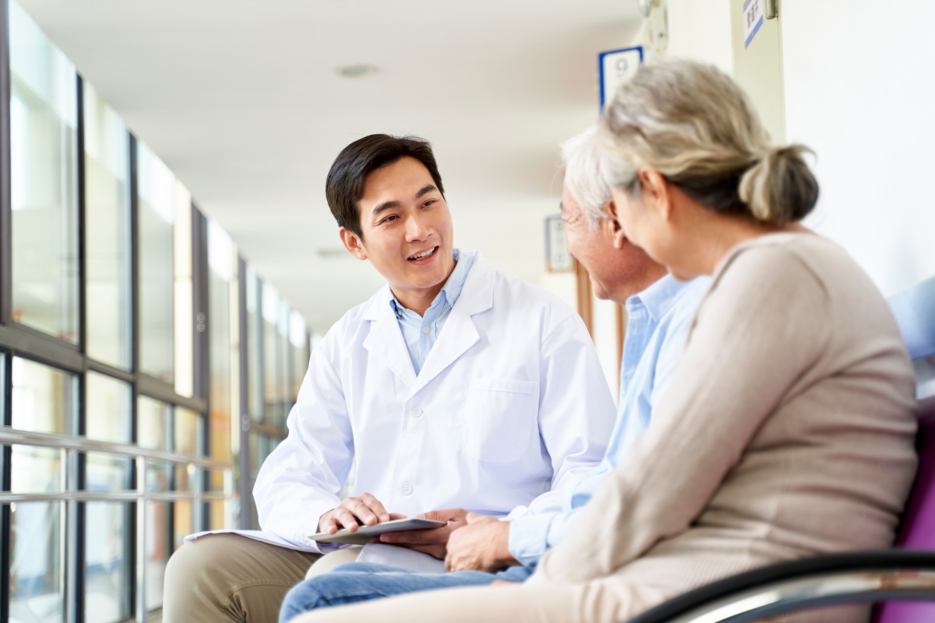 friendly young asian doctor talking to old couple in hospital hallway