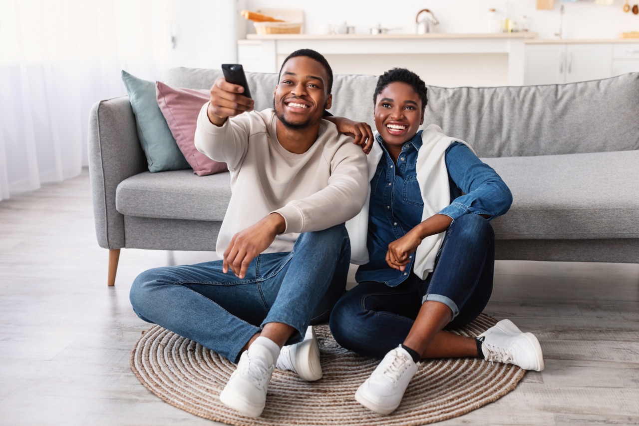 Leisure And Entertainment. Smiling African American couple watching TV show or film, guy holding remote control. Young man and woman enjoying free time sitting on floor carpet at home in living room.