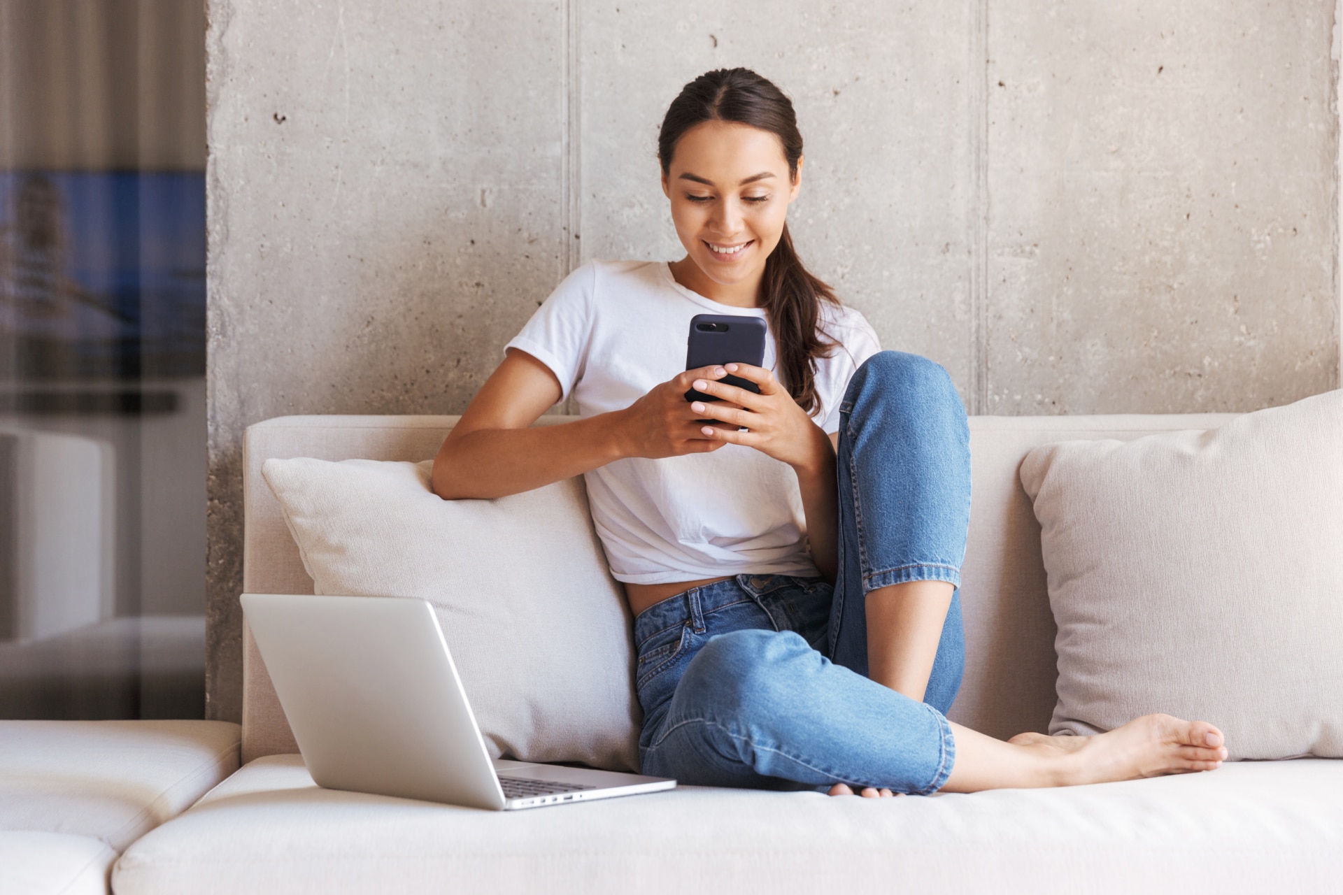 Happy young asian woman using mobile phone while sitting a couch at home with laptop computer