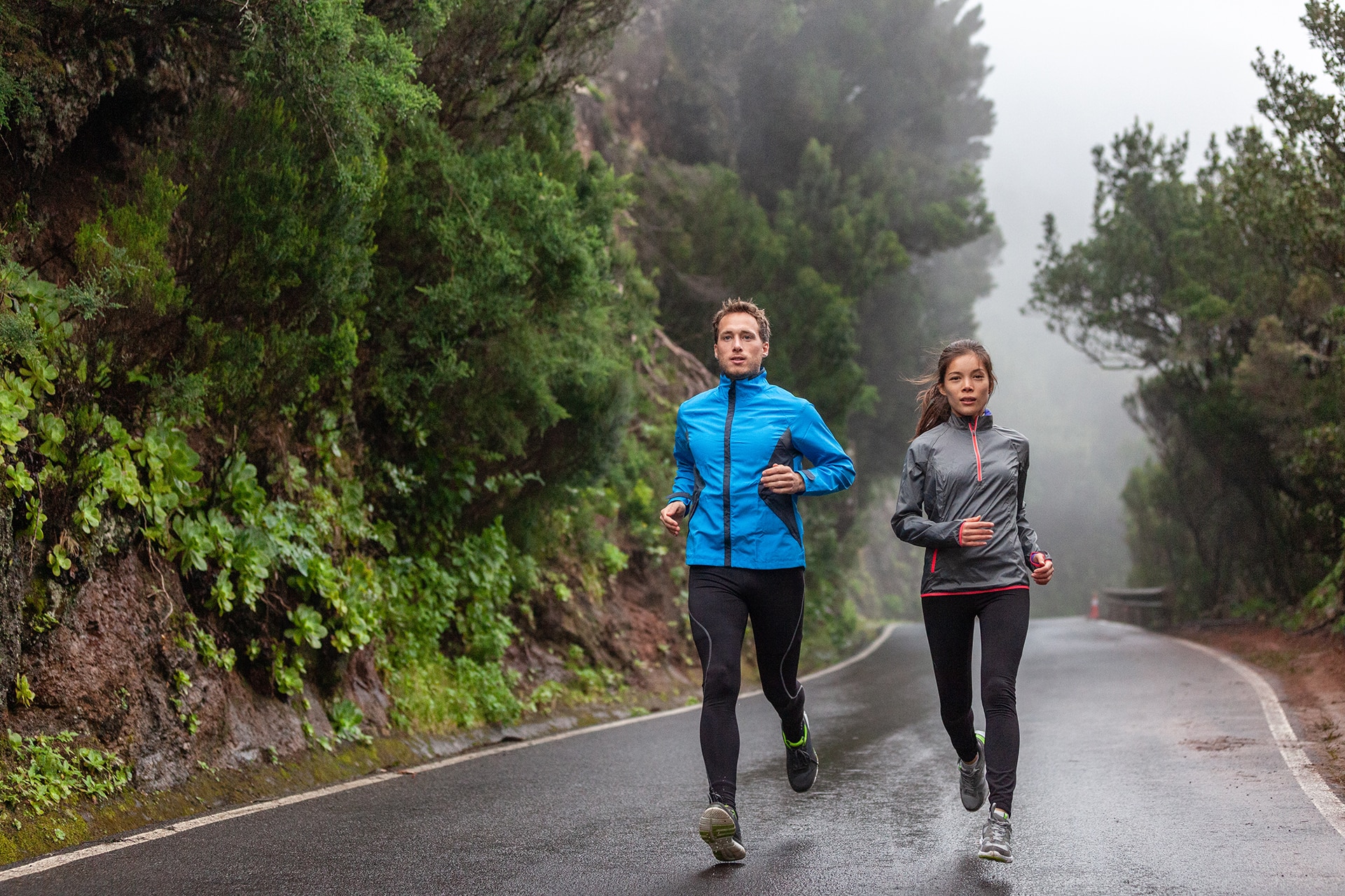 Rain run fit people training jogging in rain weather wearing cold clothing running outdoors in nature autumn season. Active couple on wet park trail jogging. Asian woman, Caucasian man athletes.