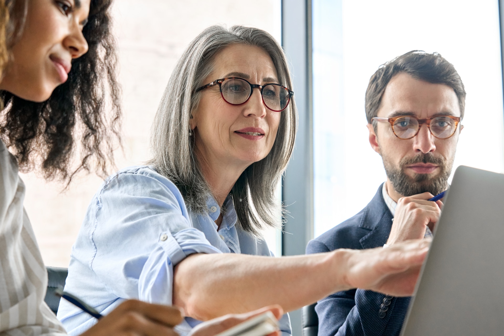 Mature older ceo businesswoman mentor in glasses negotiating growth business plan with diverse executive managers at boardroom meeting table using laptop. Multicultural team work together in office.; Shutterstock ID 2025930416; purchase_order: 1100061200; job: 600704; client: Carolin Calefi Dias; other: Phonak