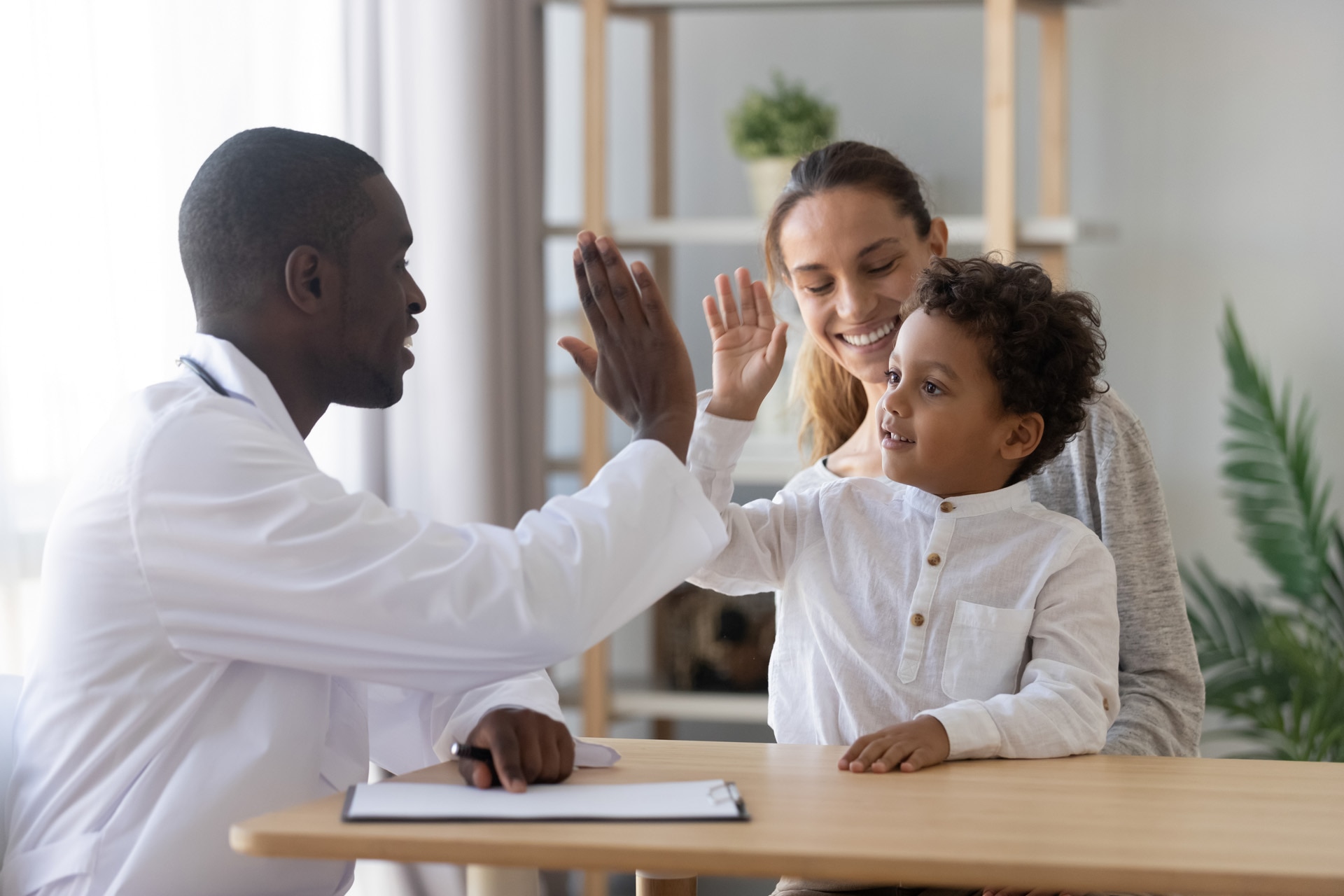 Sonriente y guapo niño mestizo choca la mano con un pediatra africano que le da la bienvenida al pequeño paciente y a su madre a la consulta; el niño confía en el doctor y celebra con él los buenos resultados médicos de su tratamiento sanitario