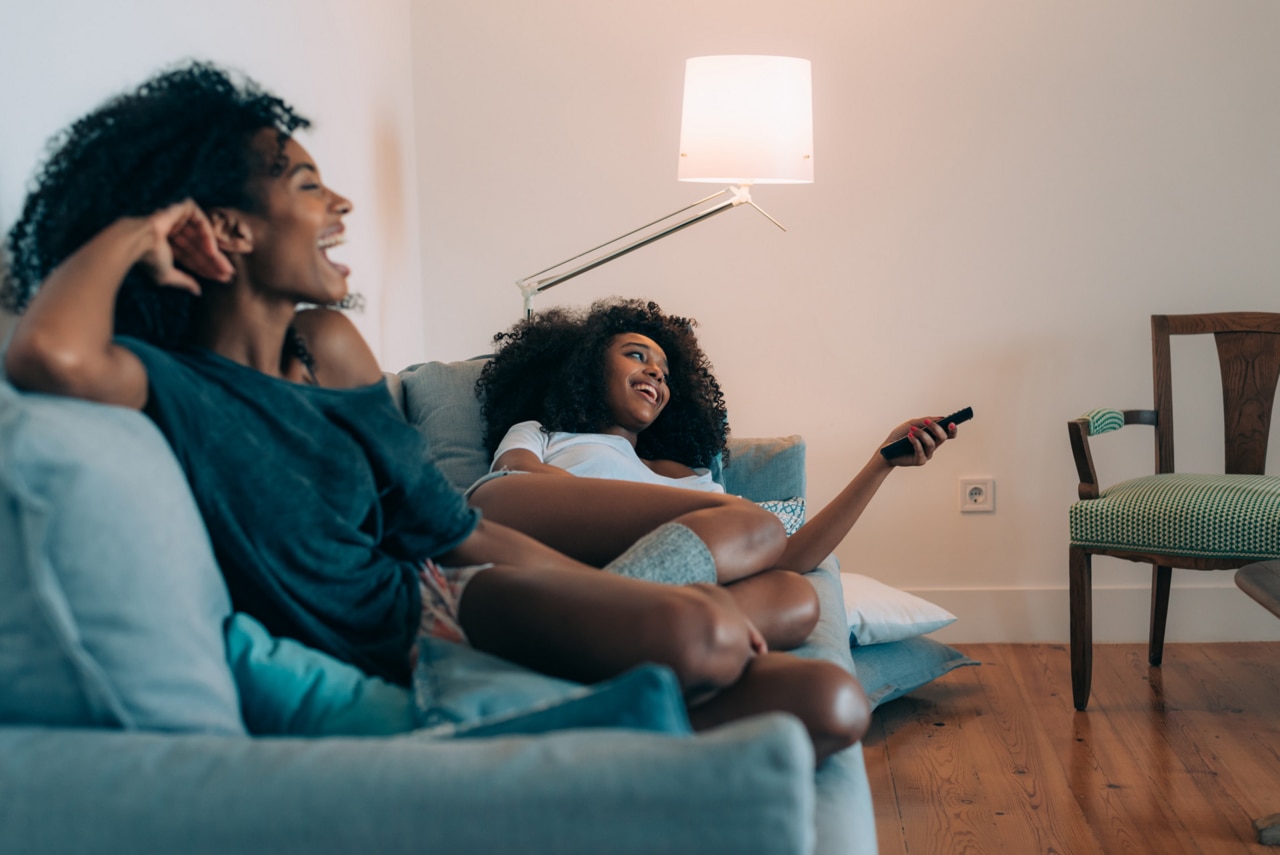Dos mujeres negras jóvenes y sonrientes recostadas en el sofá viendo la televisión 
