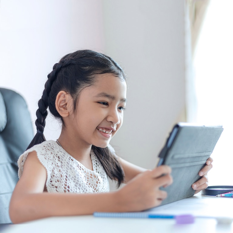 Niña de unos 7 años sonriendo y mirando algo en su tableta mientras está sentada en un escritorio.