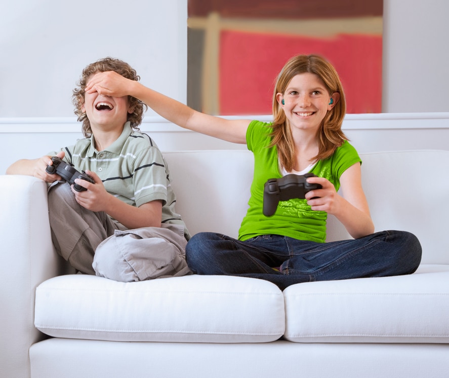 Uma menina com aparelhos auditivos e uma camiseta verde brilhante e um menino sentado em um sofá, cada um segurando um controle de videogame, envolvidos em um jogo.