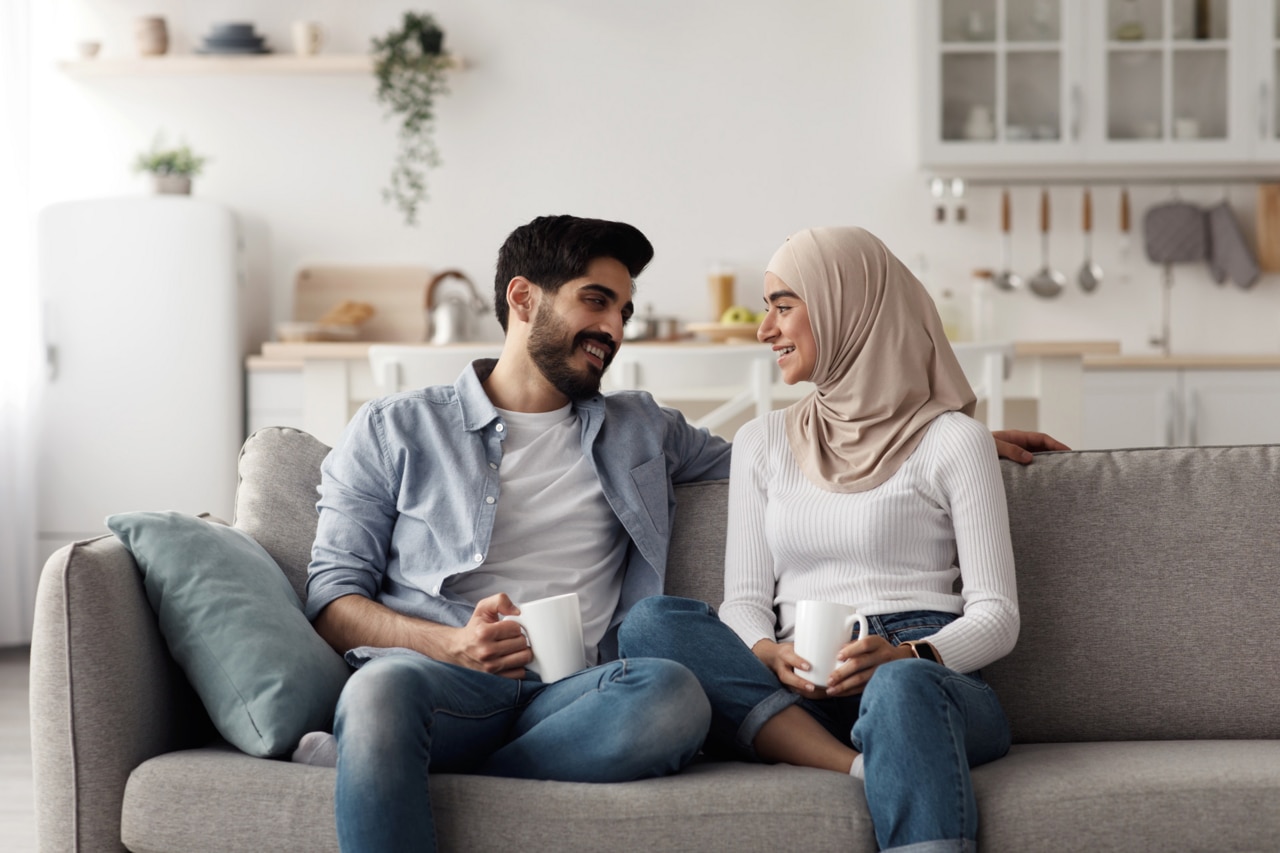 Smiling attractive young arab lady in hijab and man talking and holding cups with empty space on sofa in living room interior