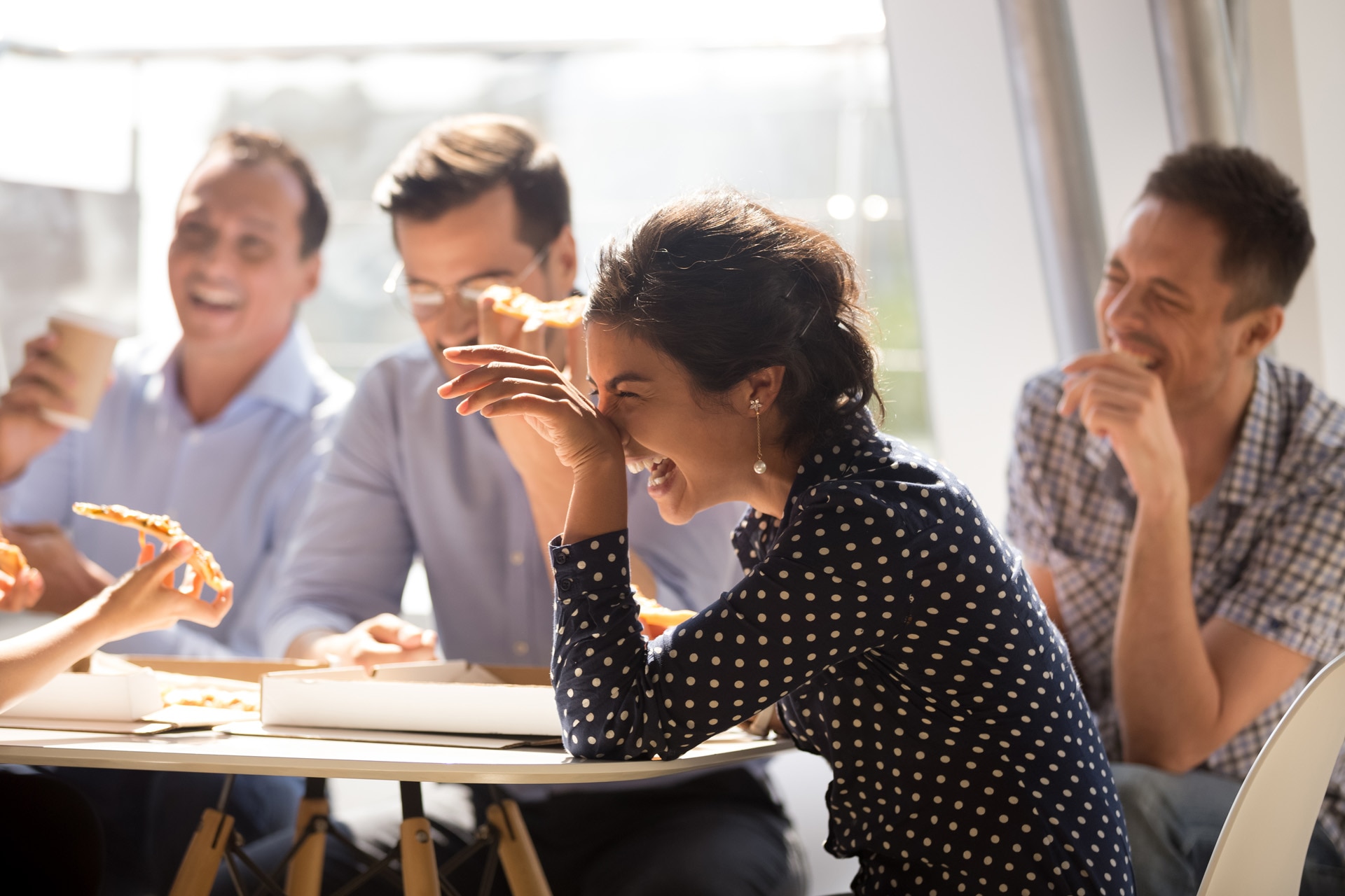 Indian woman laughing at funny joke eating pizza with diverse coworkers in office, friendly work team enjoying positive emotions and lunch together, happy colleagues staff group having fun at break; Shutterstock ID 1206996136; purchase_order: -; job: -; client: -; other: -