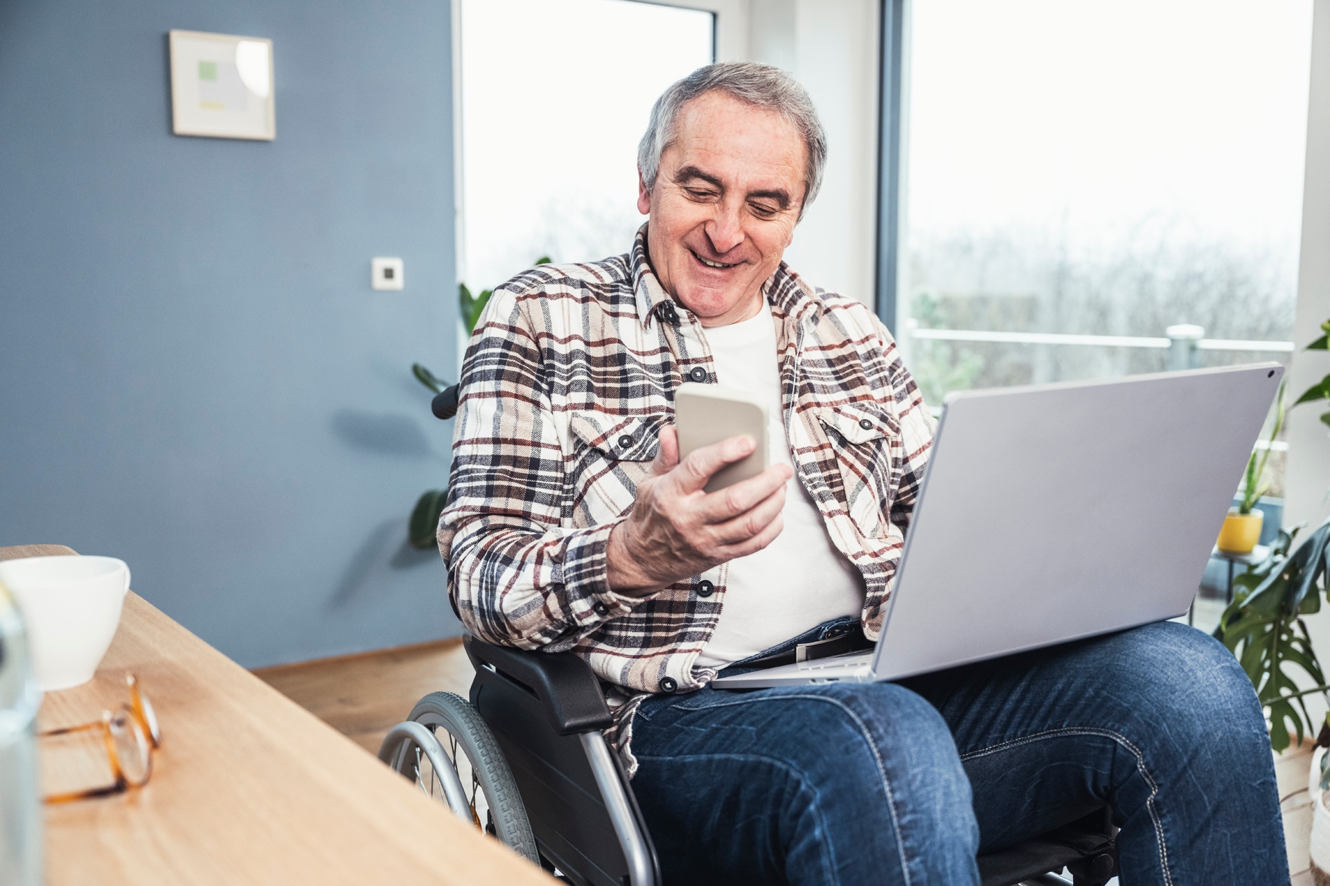Homme en situation de handicap souriant avec ordinateur portable en train d’utiliser un téléphone alors qu’il est assis dans un fauteuil roulant à la maison