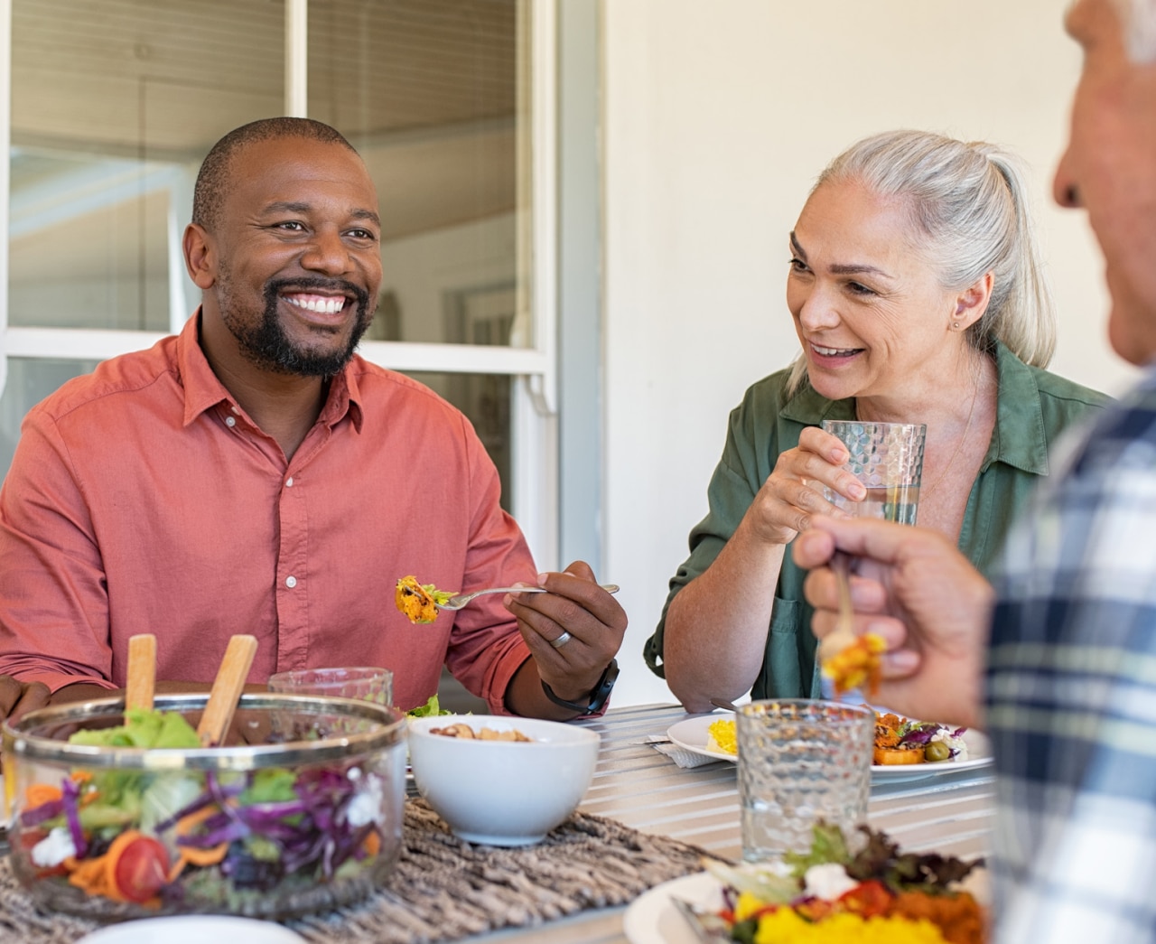 Amis heureux et souriants déjeunant ensemble dans une maison. Adultes multiethniques fêtant une heureuse occasion en mangeant de la nourriture saine. Conversation entre un couple âgé et un couple africain lors d’un repas.