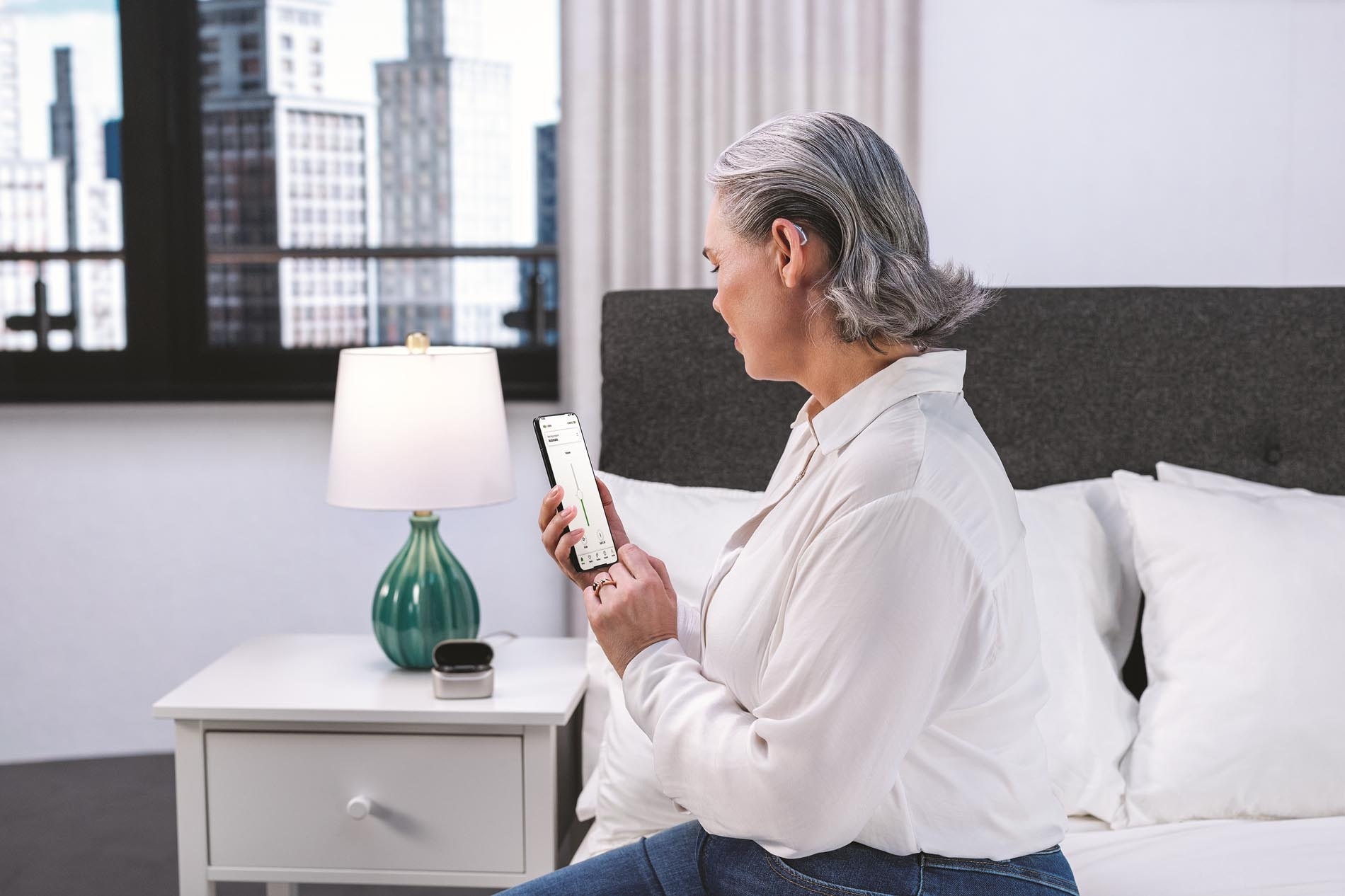 Eine ältere Frau sitzt auf einem Bett in einem hell beleuchteten Zimmer und schaut auf Ihr Smartphone