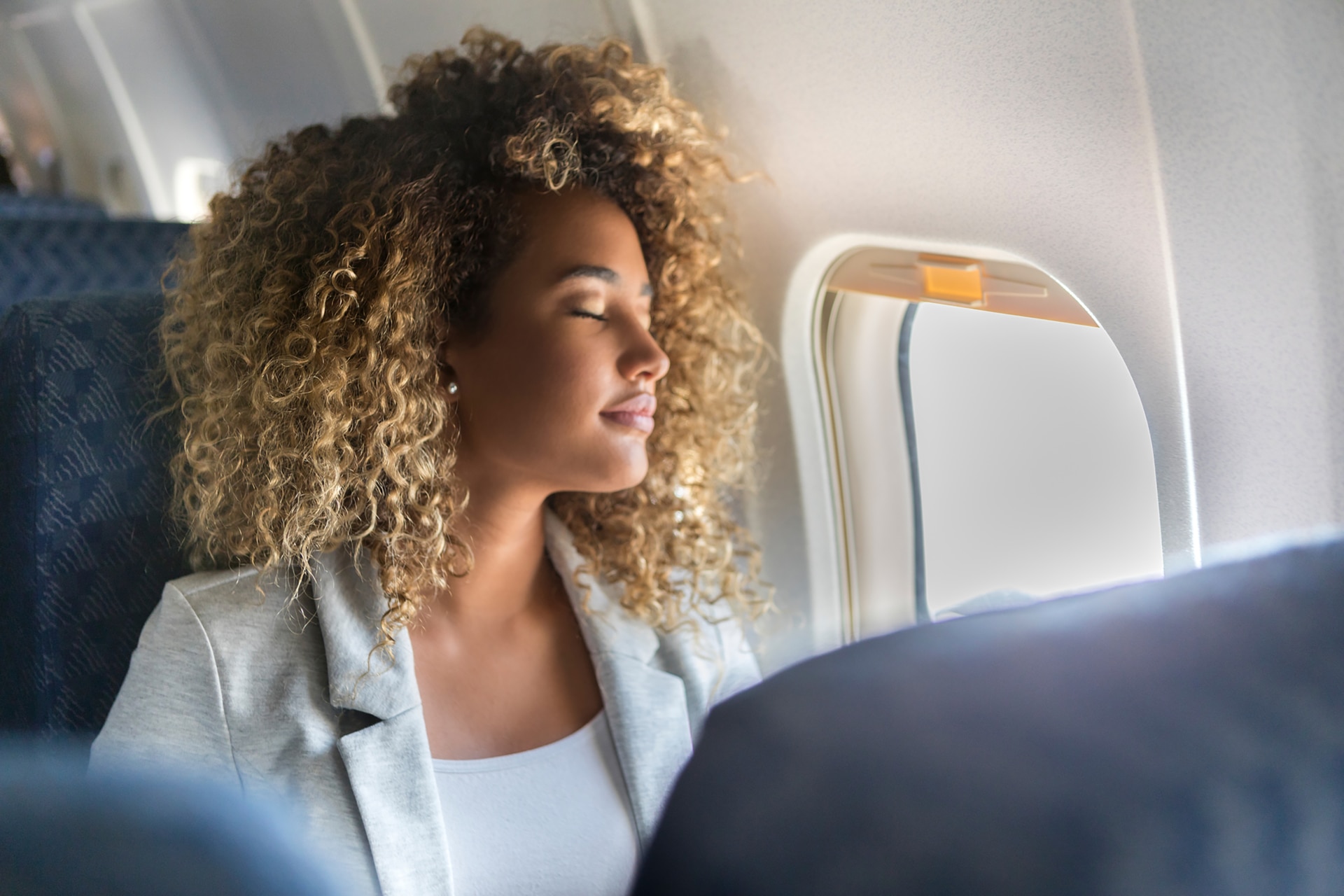 1042602524 Eine junge Frau sitzt am Fensterplatz eines Linienflugzeugs und lehnt sich mit geschlossenen Augen zurück.