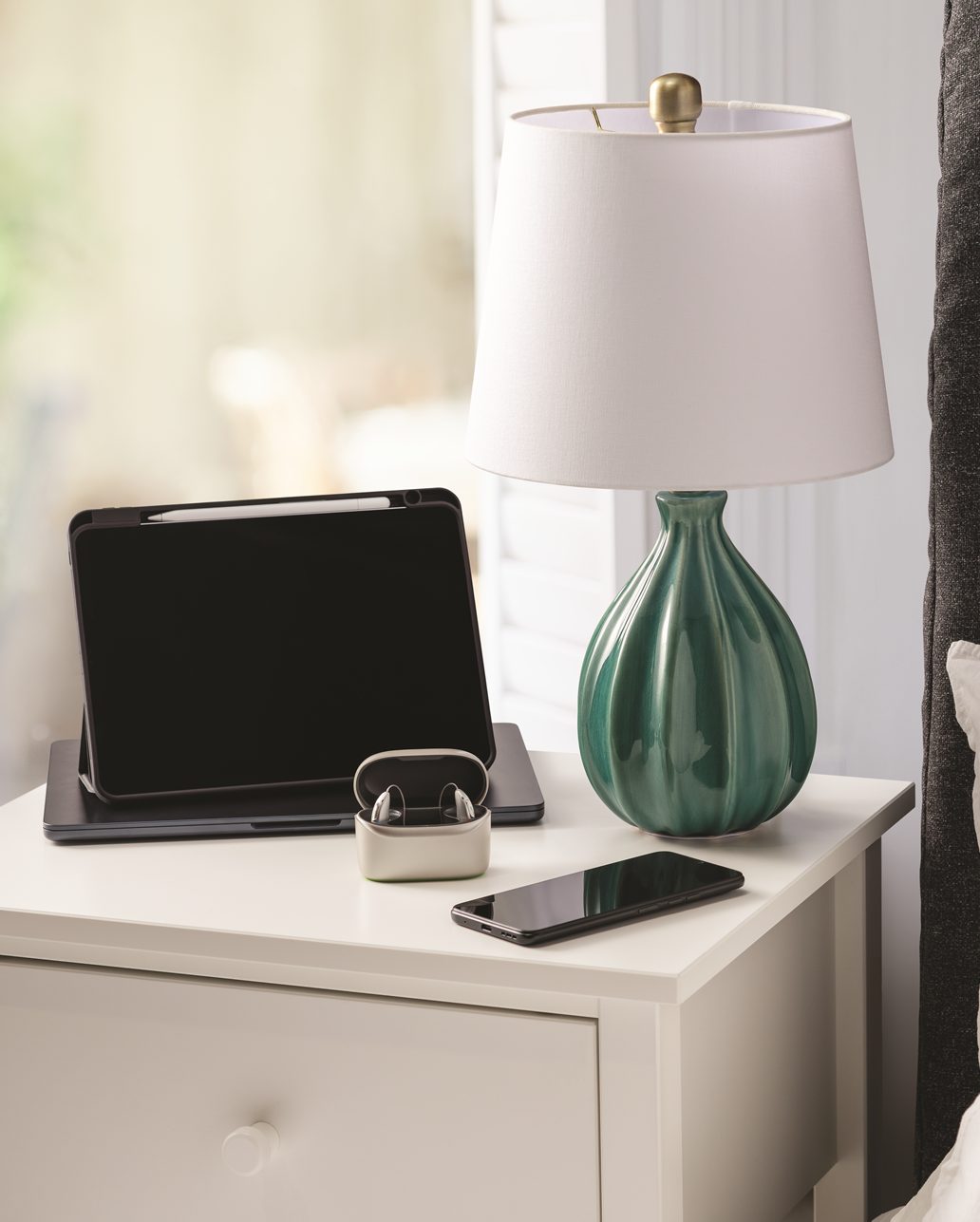 Une paire d’aides auditives dans leur chargeur sur une table de nuit, à côté d’une lampe, d’un téléphone portable et d’une tablette