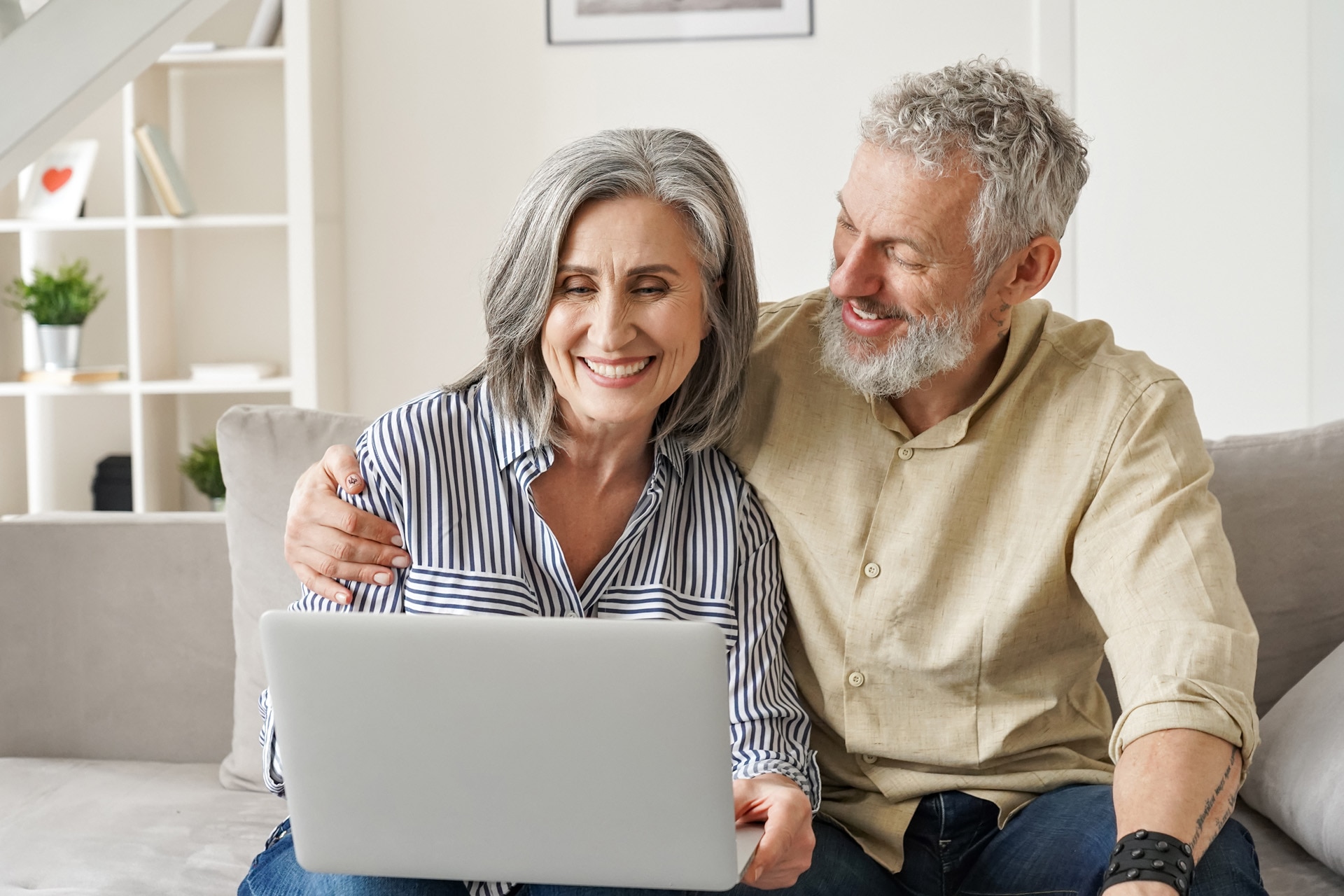 Couple de personnes d’âge moyen qui utilisent un ordinateur portable sur un canapé pour faire des achats en ligne sur un site internet. Homme et femme âgés en train de regarder un ordinateur alors qu’ils font une recherche internet pour souscrire une assurance depuis chez eux.