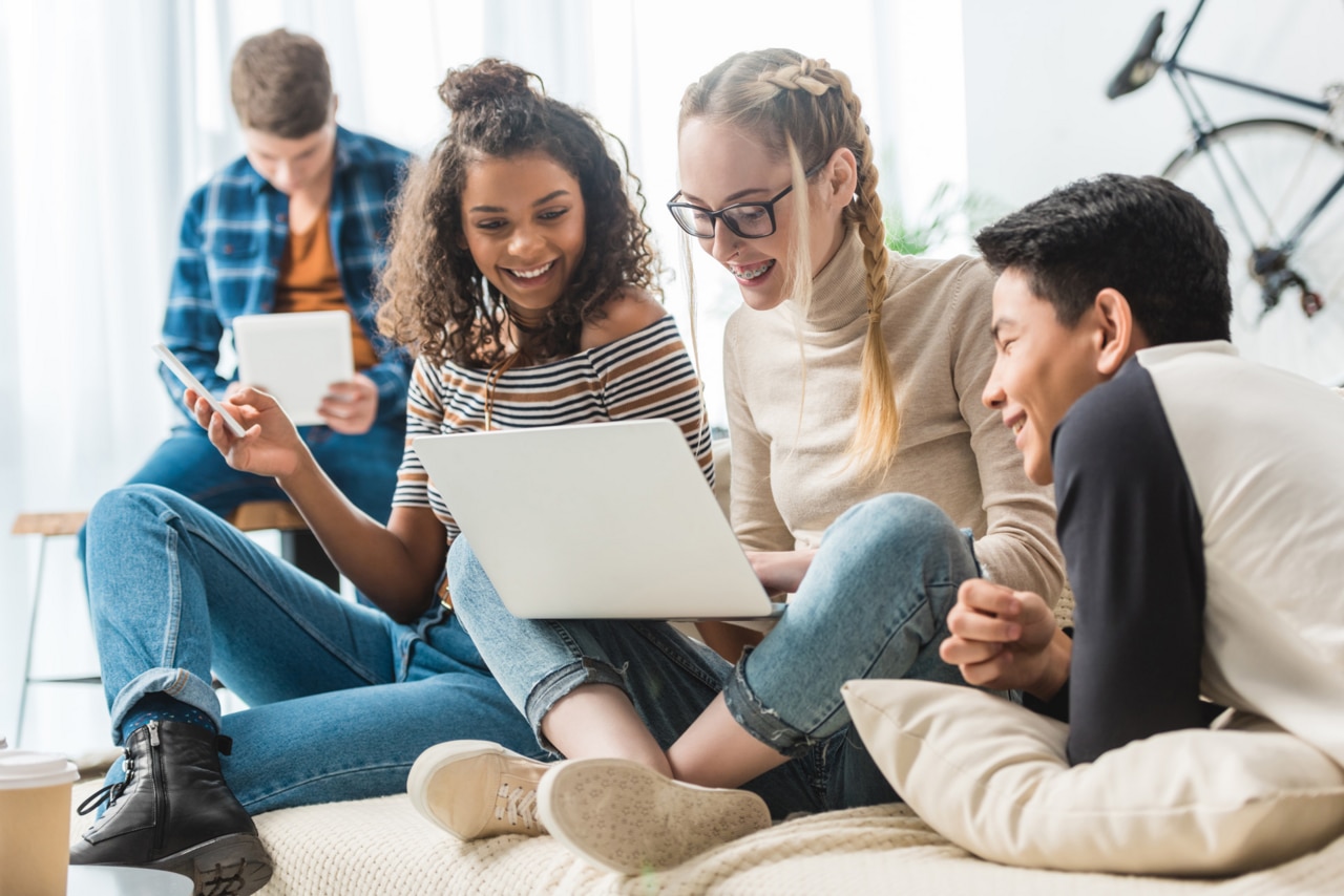 adolescents multiculturels heureux regardant un ordinateur portable et assis sur un canapé