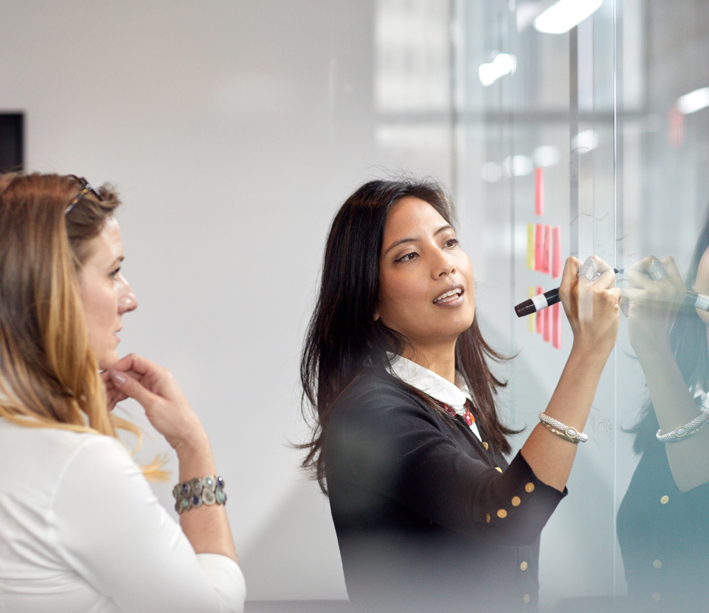 Businesswoman looking at female colleague writing on adhesive notes in office