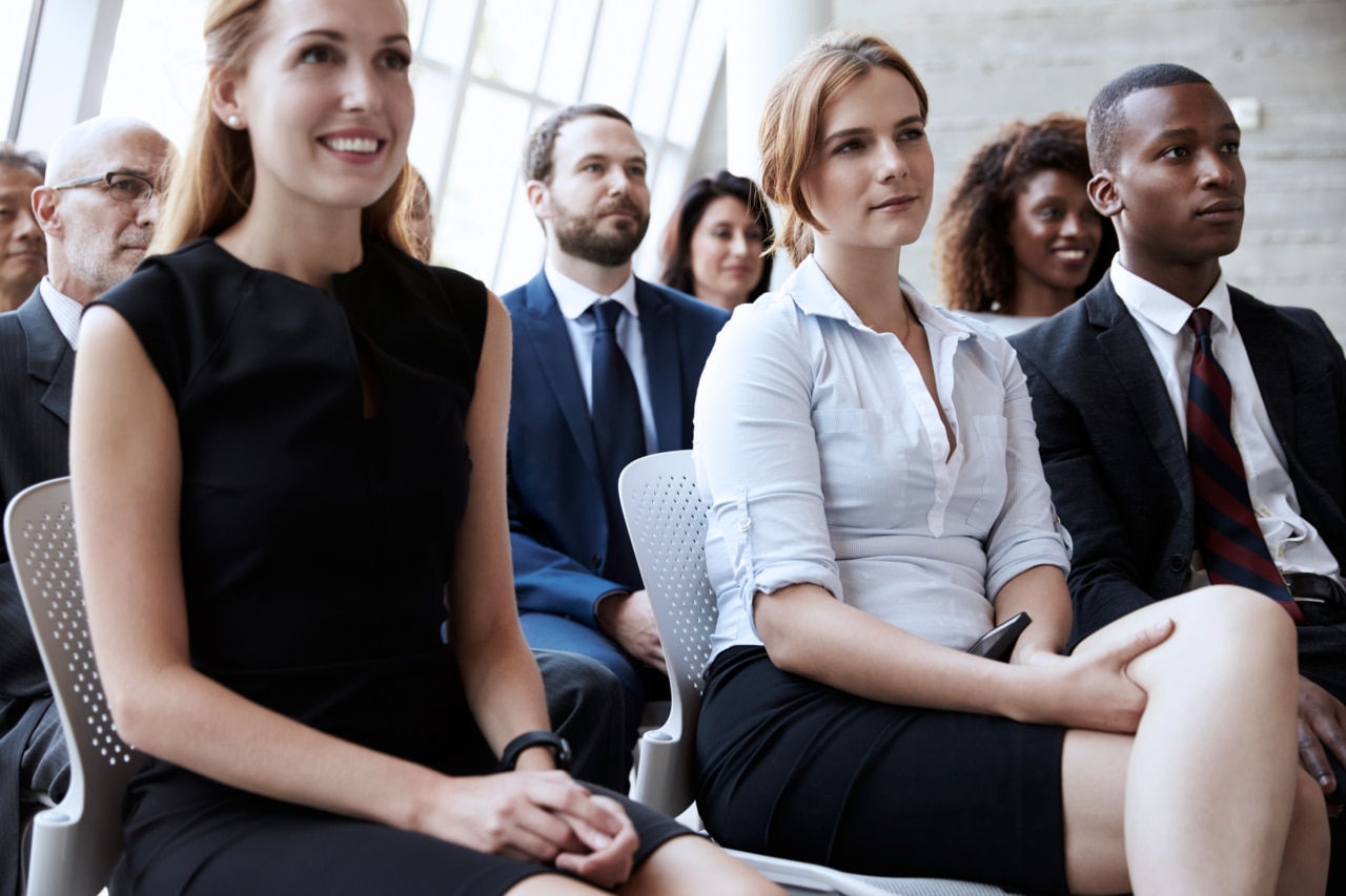 A man seated rows behind the speaker sits in a crowded auditorium watching a presentation.