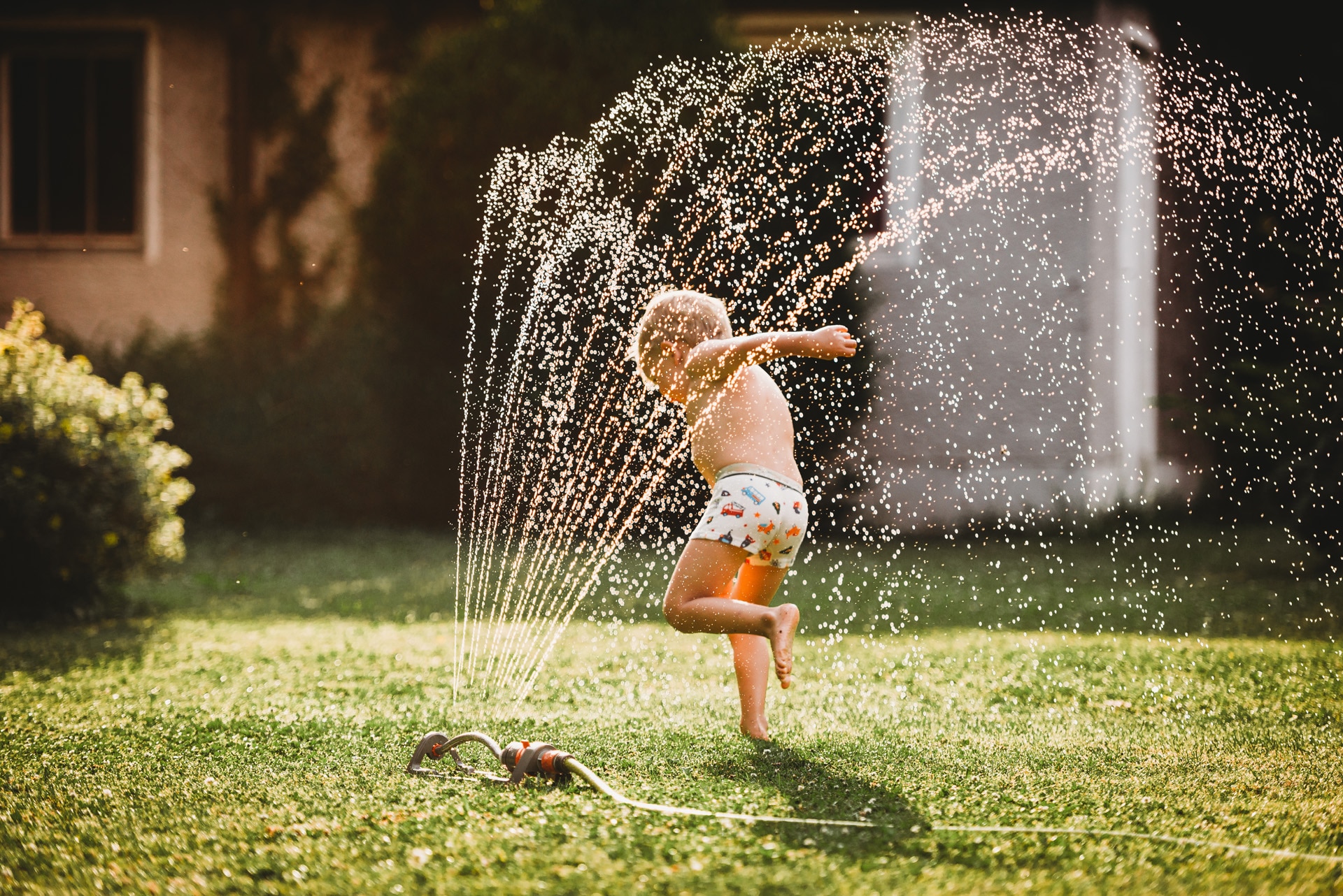 Kleiner Junge läuft unter dem Wasserstrahl eines Rasensprengers im Garten.