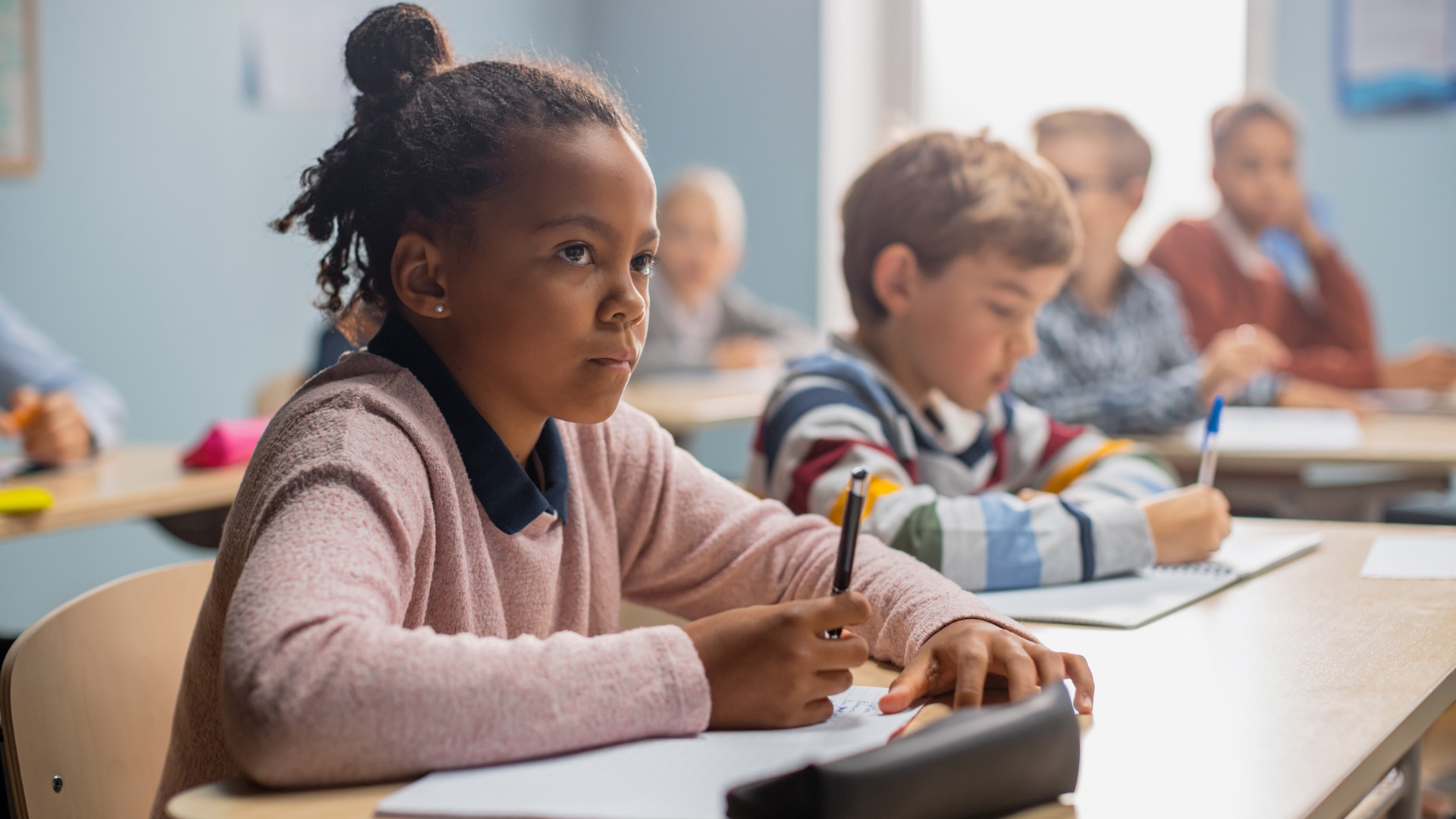 In Elementary School Classroom Brilliant Black Girl Writes in Exercise Notebook, Taking Test and Writing Exam. Junior Classroom with Diverse Group of Children Working Diligently and Learning