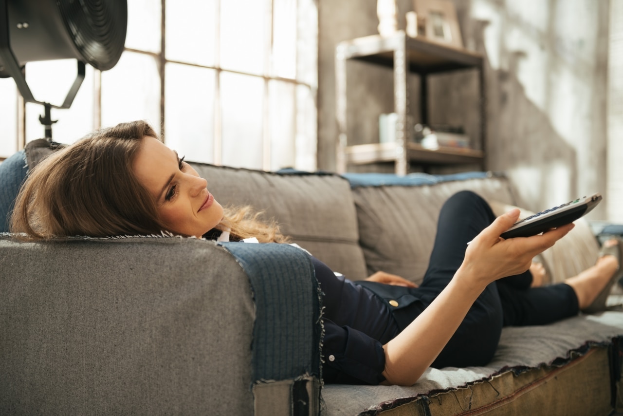 Afslappet ung kvinde ligger på en sofa og ser TV i en loftslejlighed. Smart bydekoration i loftet og vindue.