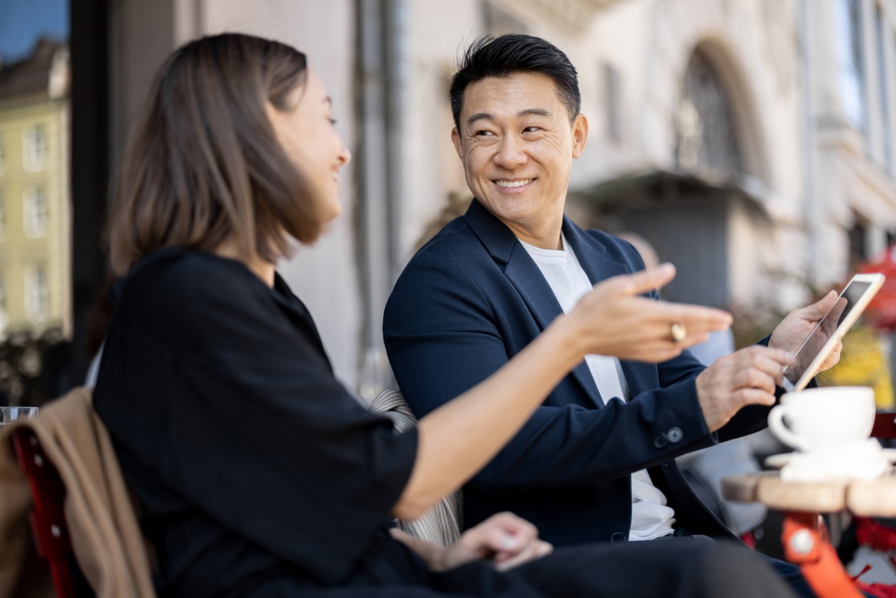 Caucasian woman and asian man having a business conversation while sitting at outdoor cafe. Concept of remote and freelance work while resting. Idea of teamwork and business cooperation; Shutterstock ID 2074091051; purchase_order: -; job: -; client: -; other: -