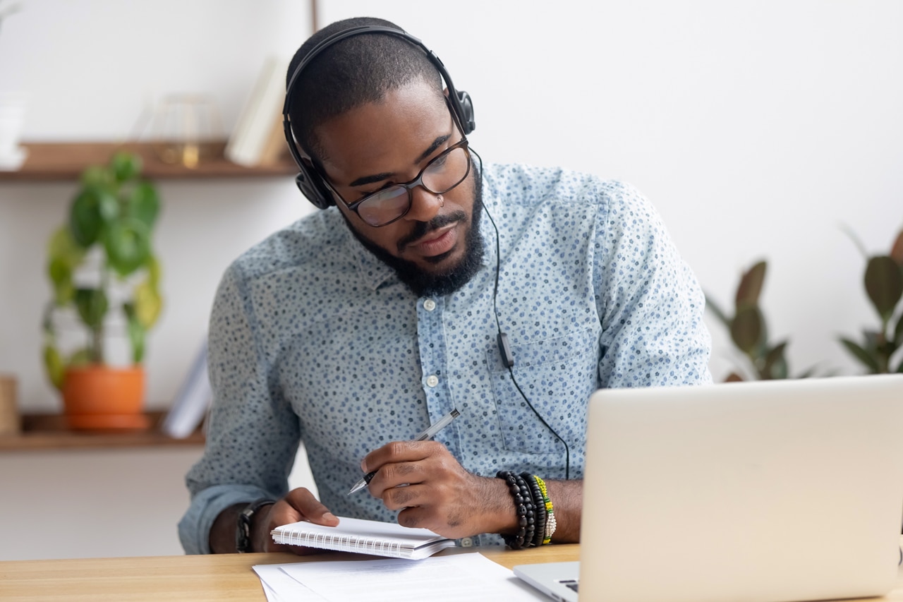 Hombre de negocios africano enfocado con audífonos escribiendo notas en un cuaderno viendo un curso de video de seminario web, estudiante negro serio mirando un estudio de conferencias escuchando una computadora portátil en línea sobre aprendizaje electrónico por computadora;
