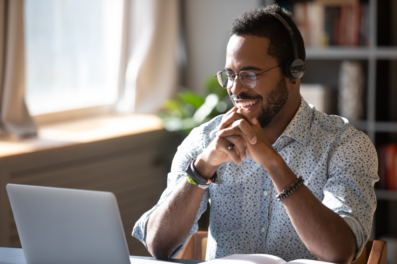 Feliz hombre afroamericano milenario con gafas y audífonos, disfrutando viendo un seminario web educativo en una computadora portátil. Sonriente joven empresario de raza mixta realizando videollamadas con socios de clientes.