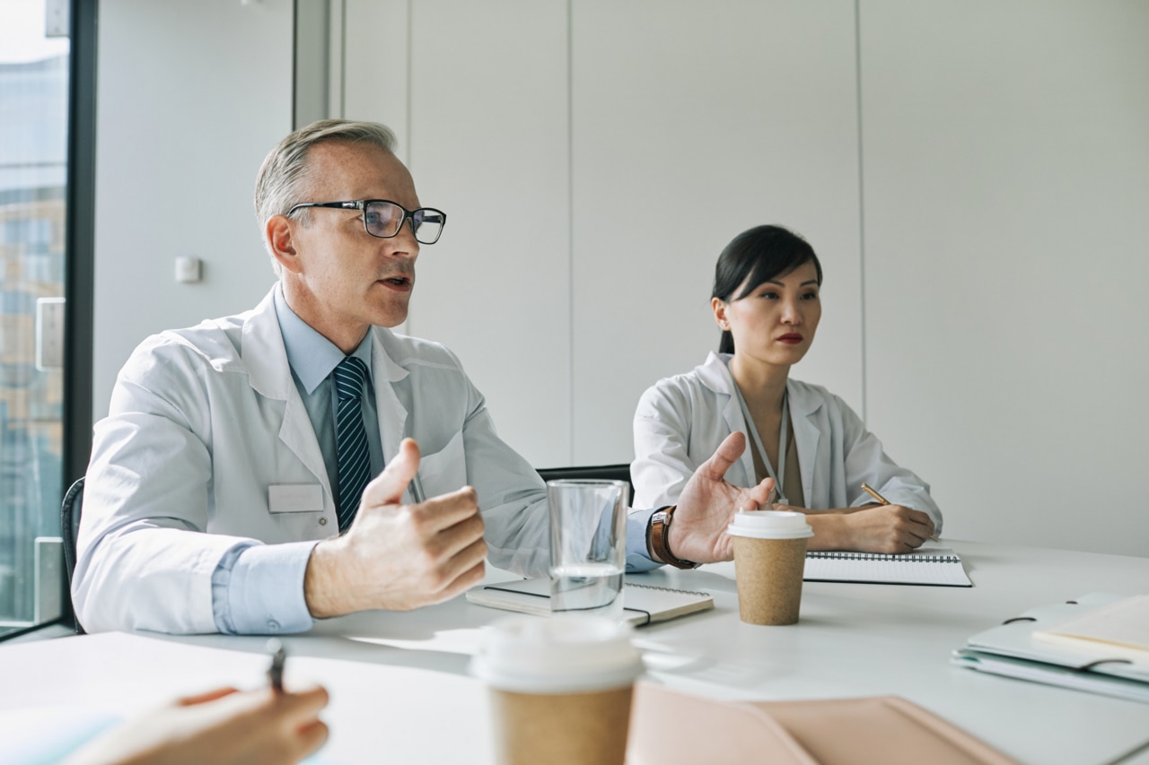 Portrait of mature doctor sitting at meeting table in conference room while speaking during medical seminar
