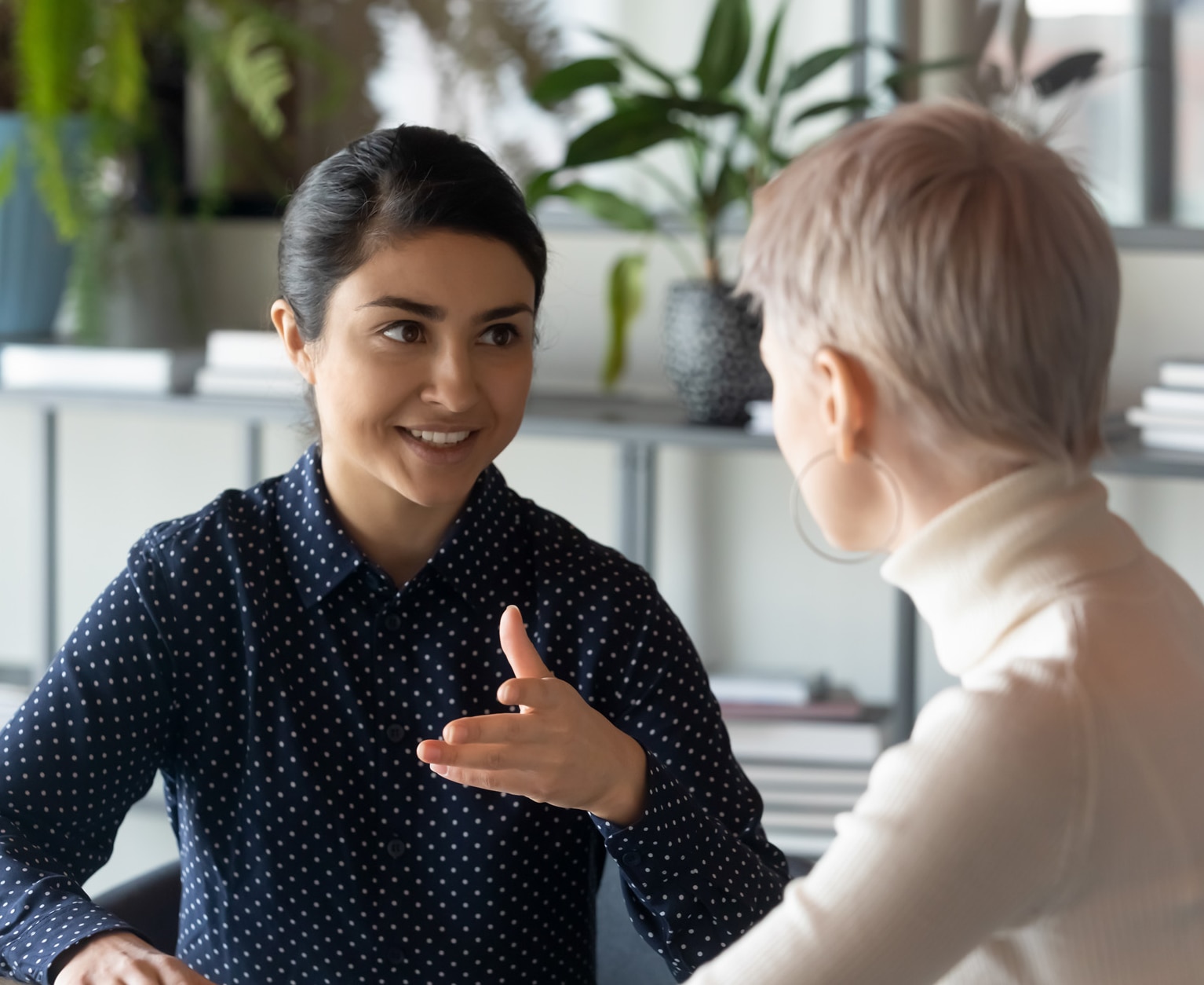 Friendly millennial indian businesswoman talking to blonde female colleague, sitting at table in office. Two young multiracial employees discussing working issues or enjoying informal conversation.; Shutterstock ID 1796961841; purchase_order: -; job: -; client: -; other: -