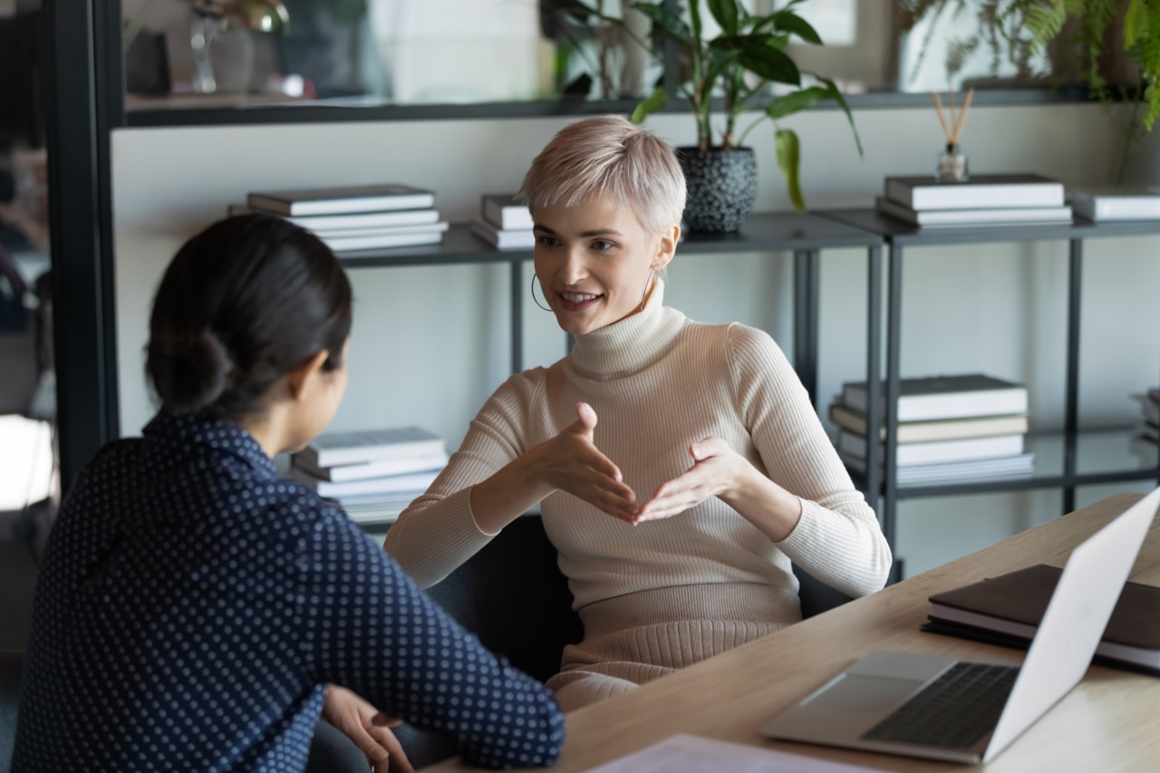 Une employée multiethnique souriante et diversifiée s'assoit au bureau pour discuter d'idées commerciales et coopérer sur un ordinateur portable. Une employée multiethnique souriante et diversifiée s'assoit au bureau pour discuter d'idées commerciales et coopérer sur un ordinateur portable.
