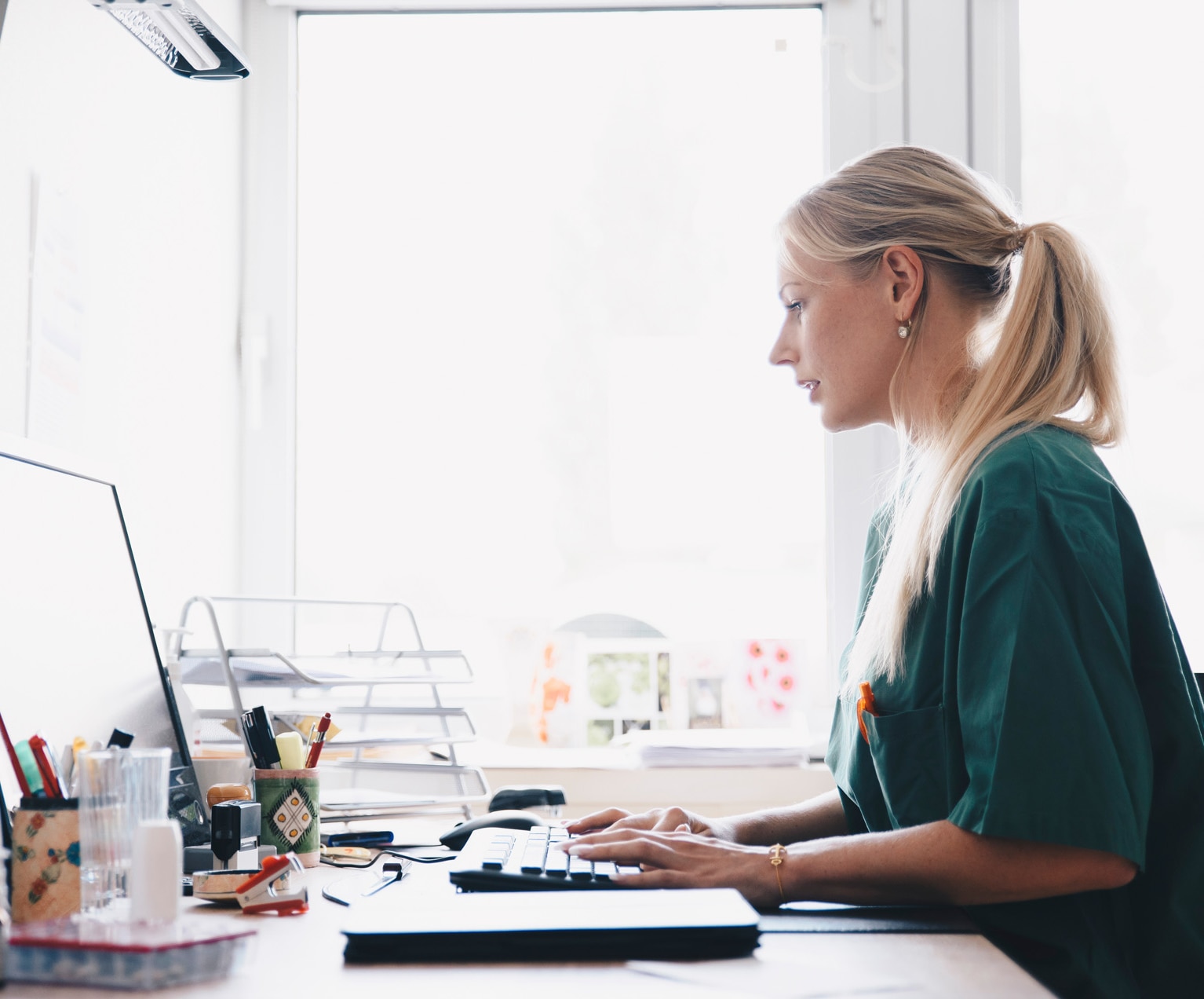 Side view of female nurse working at computer desk against window in office