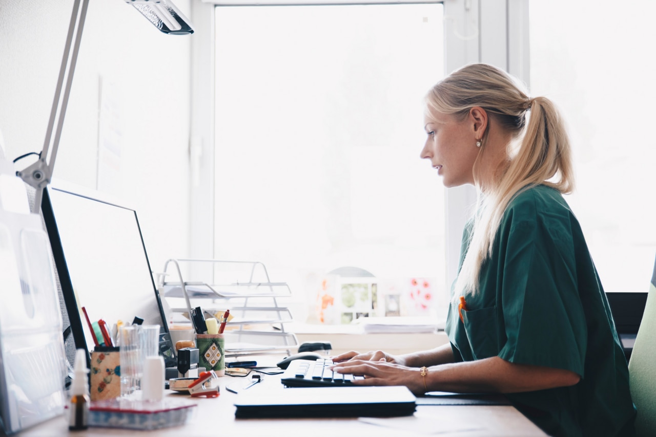 Vue latérale d'une infirmière travaillant au bureau d'ordinateur contre une fenêtre au bureau