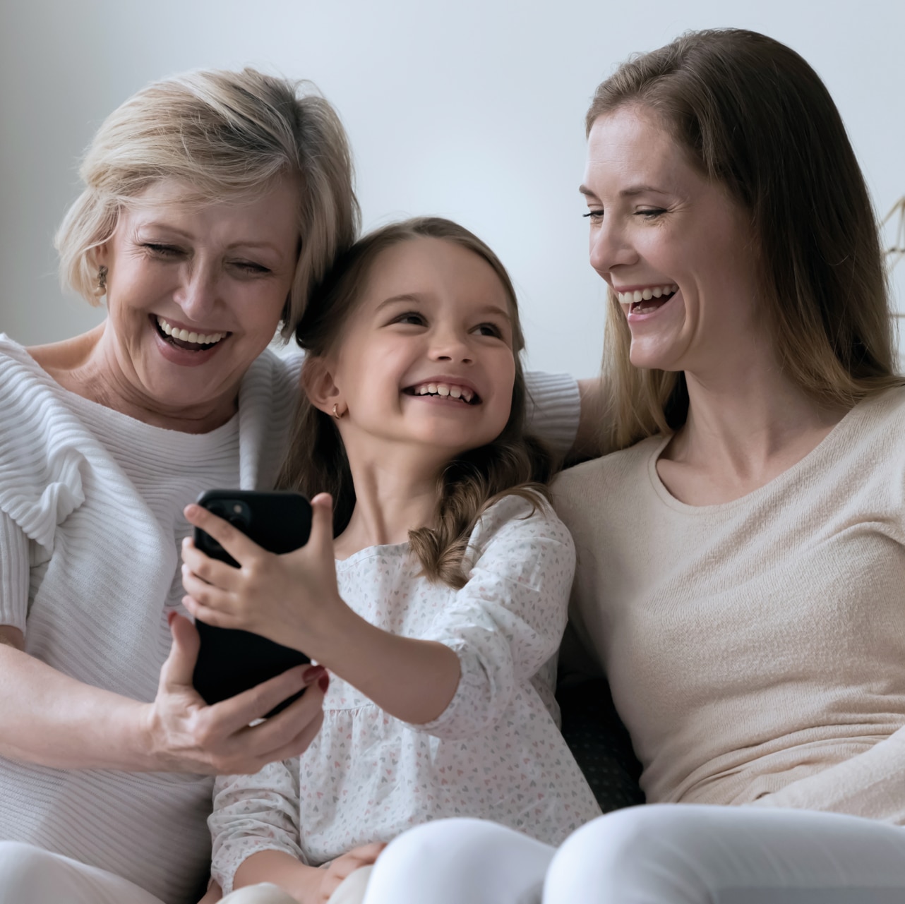 Elderly 60s granny her daughter and granddaughter laughing watch on-line videos sit on sofa using modern smart phone, feel carefree spend leisure on internet. Diverse generation using wireless tech; Shutterstock ID 2306207029; purchase_order: -; job: -; client: -; other: -