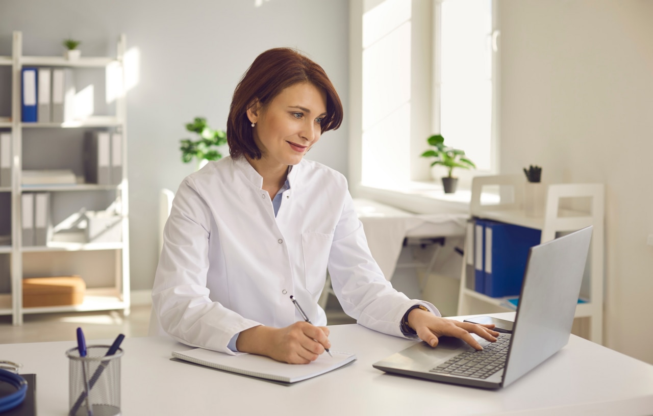 A munkahelyén egy laptopnál ülő orvosnő jegyzeteket ír, miközben online orvosi webináriumot vagy képzési szemináriumot néz. A pozitív orvos mindent megtesz a kiváló minőségű orvosi ellátás érdekében
