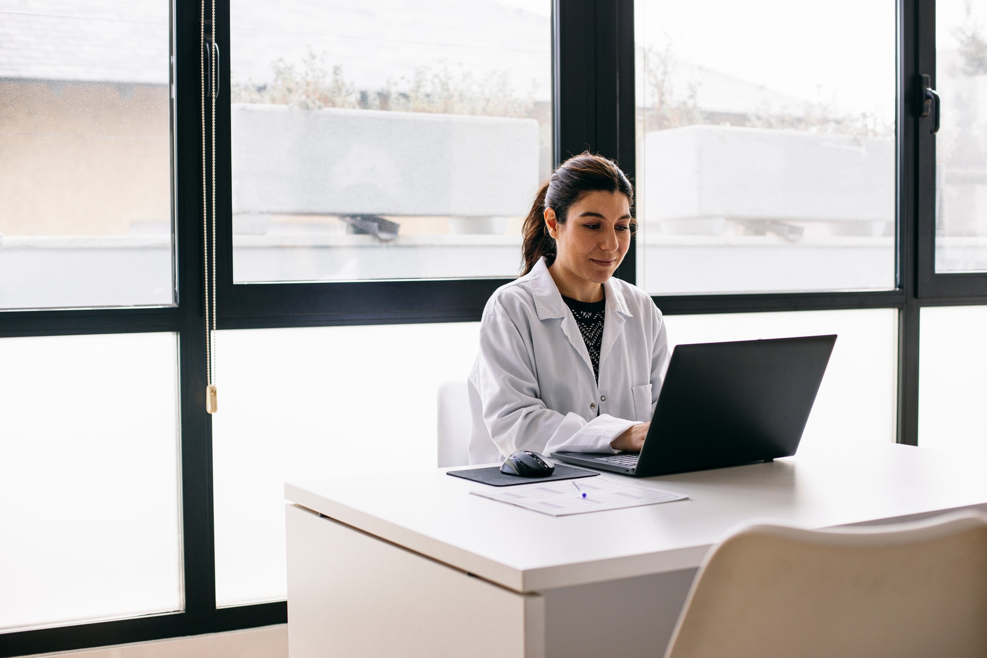 Laptopot használó orvosnő ül az íróasztalánál az orvosi rendelőben.
