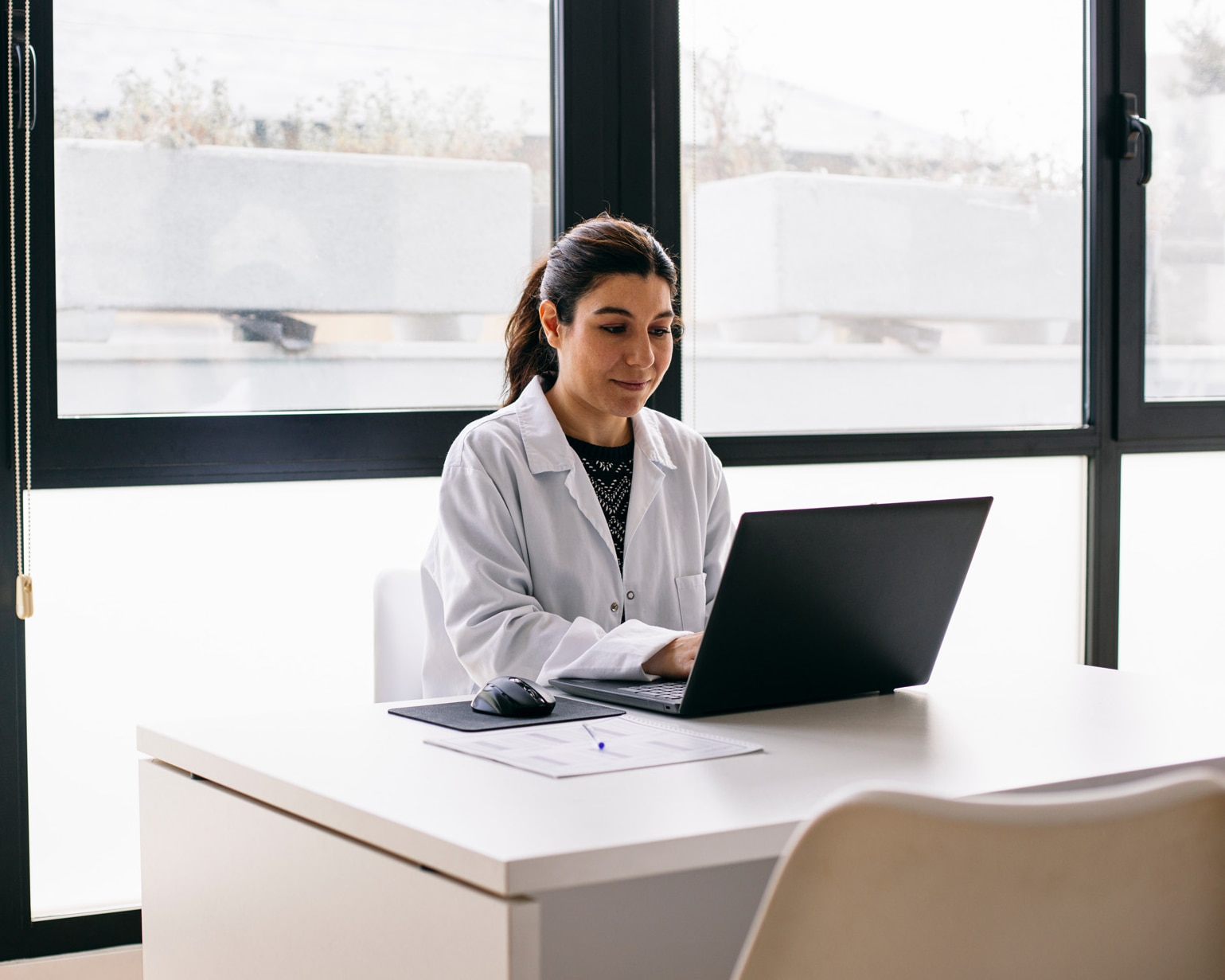 Laptopot használó orvosnő ül az íróasztalánál az orvosi rendelőben.